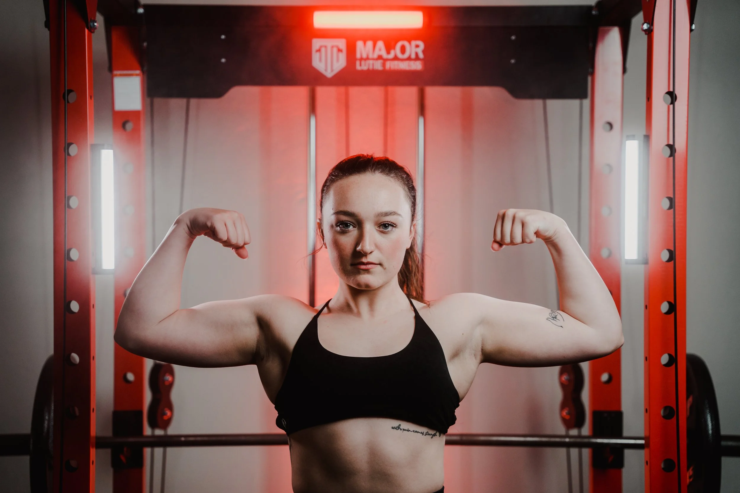 A woman in a gym flexes her biceps in front of red fitness equipment.