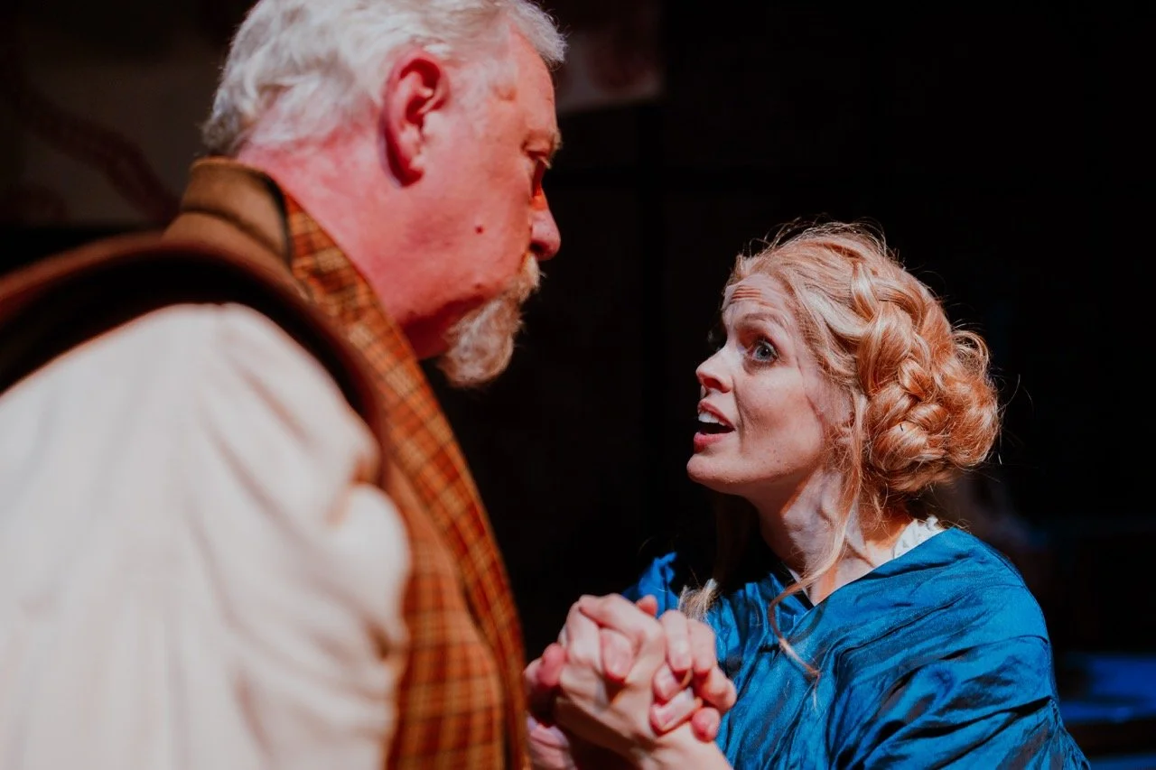 A woman with braided blonde hair and a blue shawl holds hands with an older man with white hair and a plaid scarf, engaging in an emotional conversation on stage.