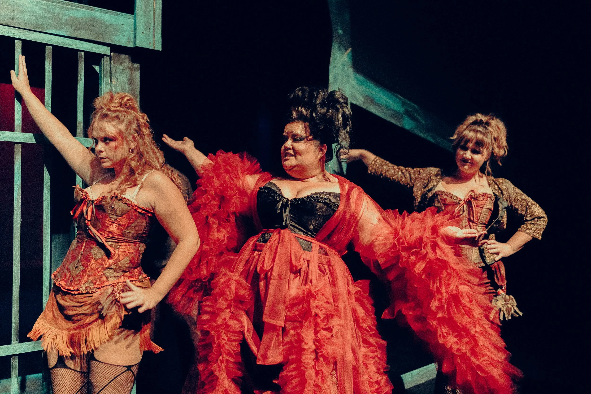 Three women in theatrical costumes, one in a red ruffled dress, perform on stage.