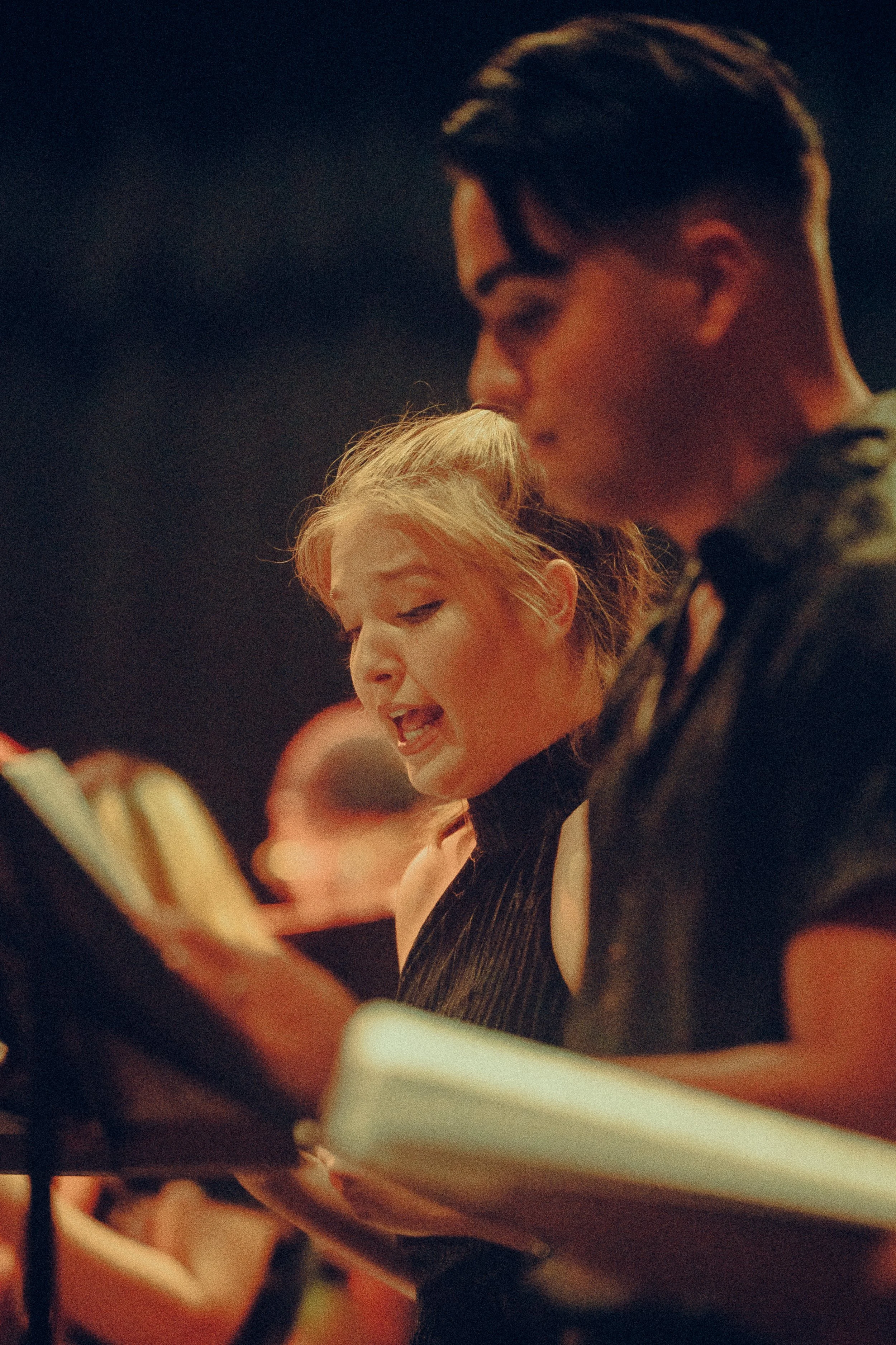 Two people, a man and a woman, are singing on stage while reading from sheet music. The setting is dimly lit, suggesting a rehearsal or performance.