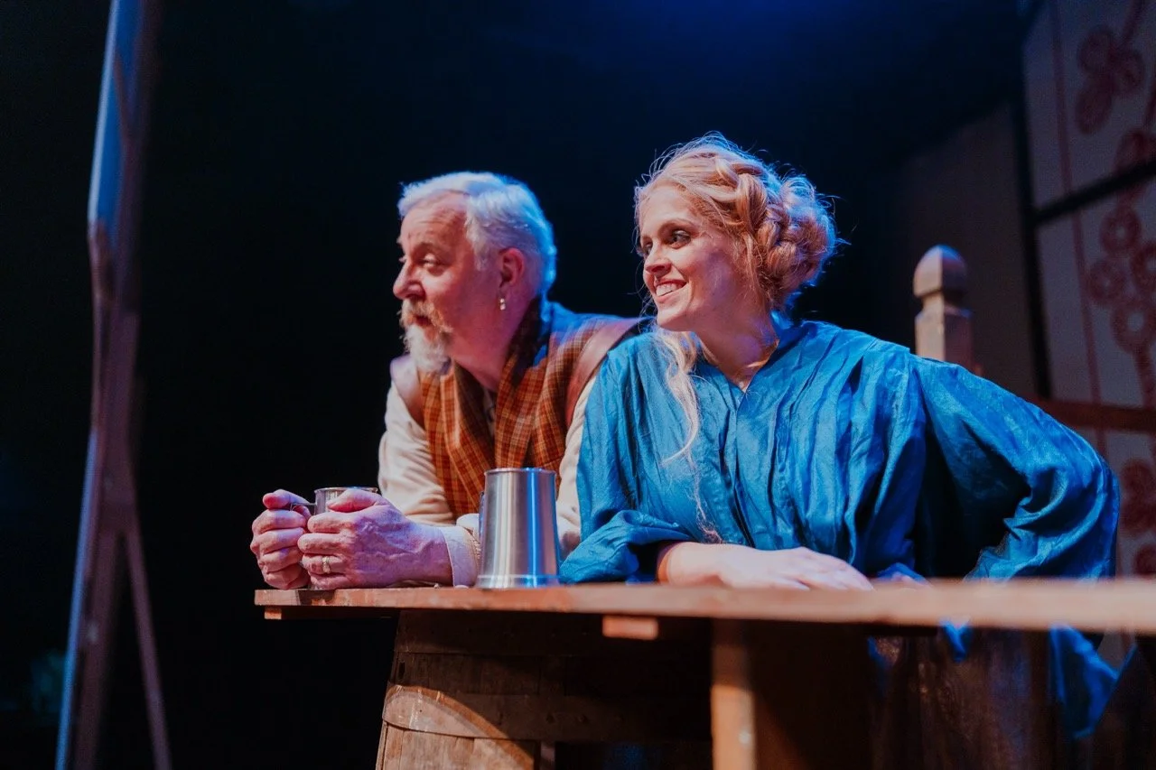 Two performers in historical costumes, a man with a beard and earring, and a woman with braided hair, leaning on a wooden surface during a stage performance.