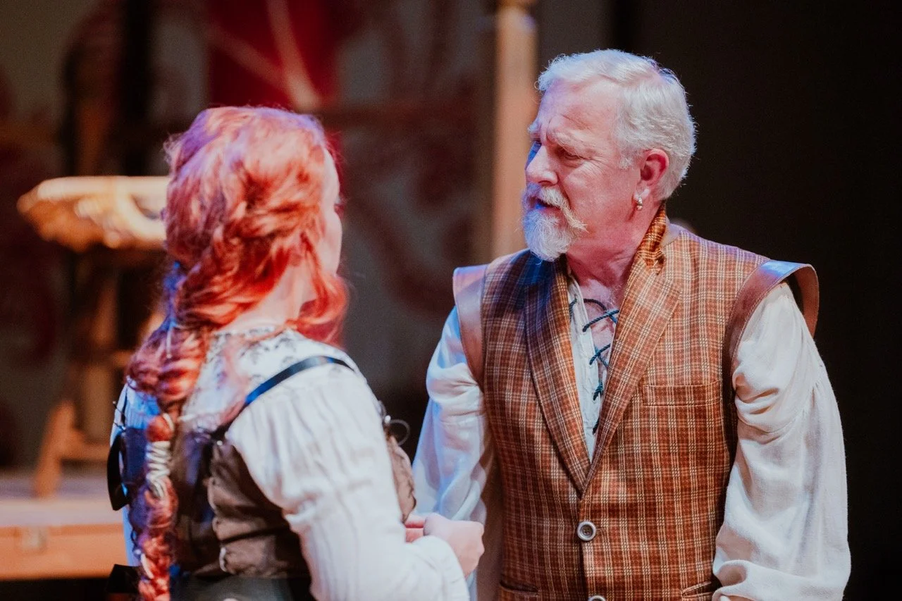 Two actors in historical costumes on stage, engaged in conversation. One has red hair in a braid, the other has white hair and a plaid vest.