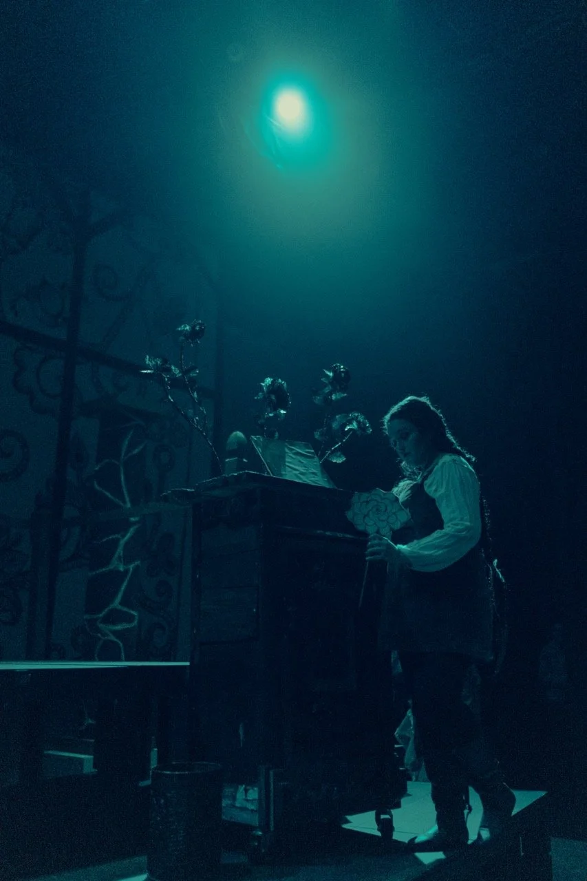 Person on stage under blue light holding a prop flower with a backdrop of plants and patterns.