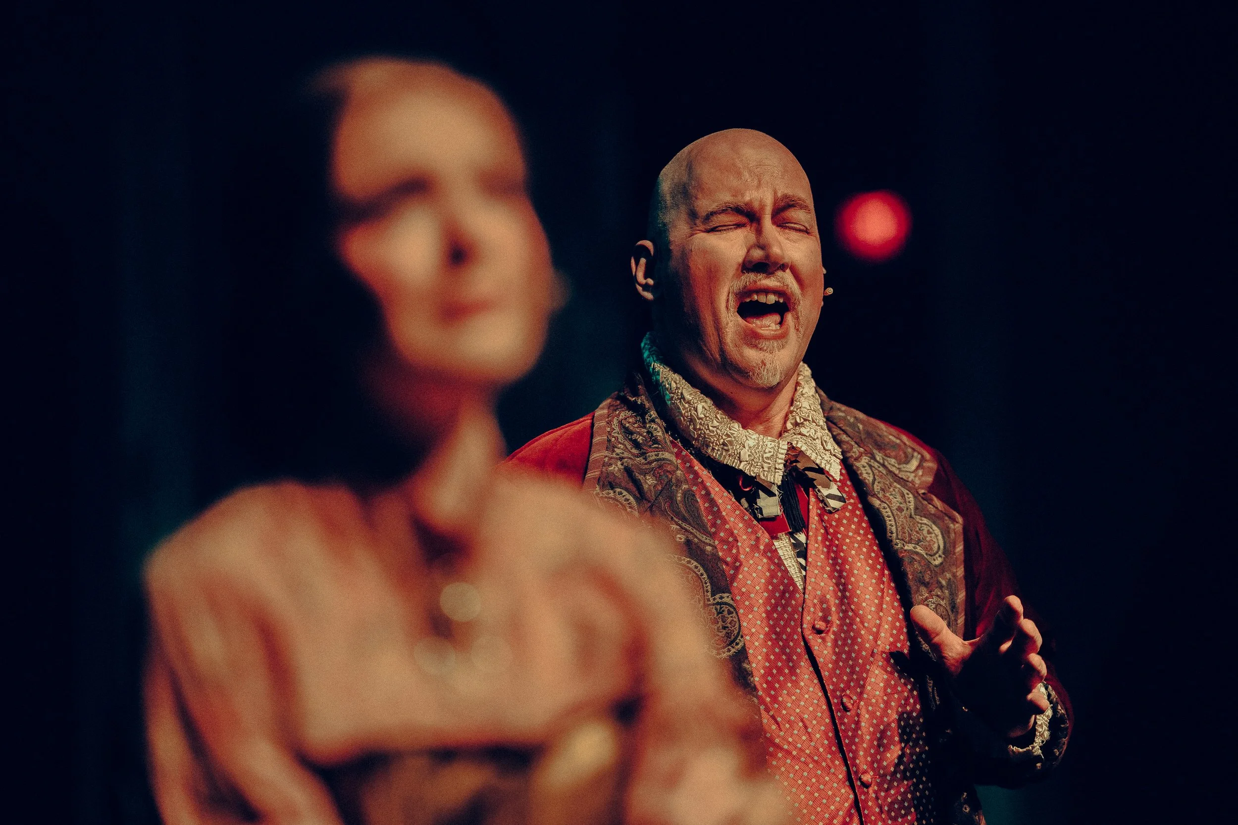 Man passionately singing with blurred figure in foreground, wearing period costume, on stage with dramatic lighting.