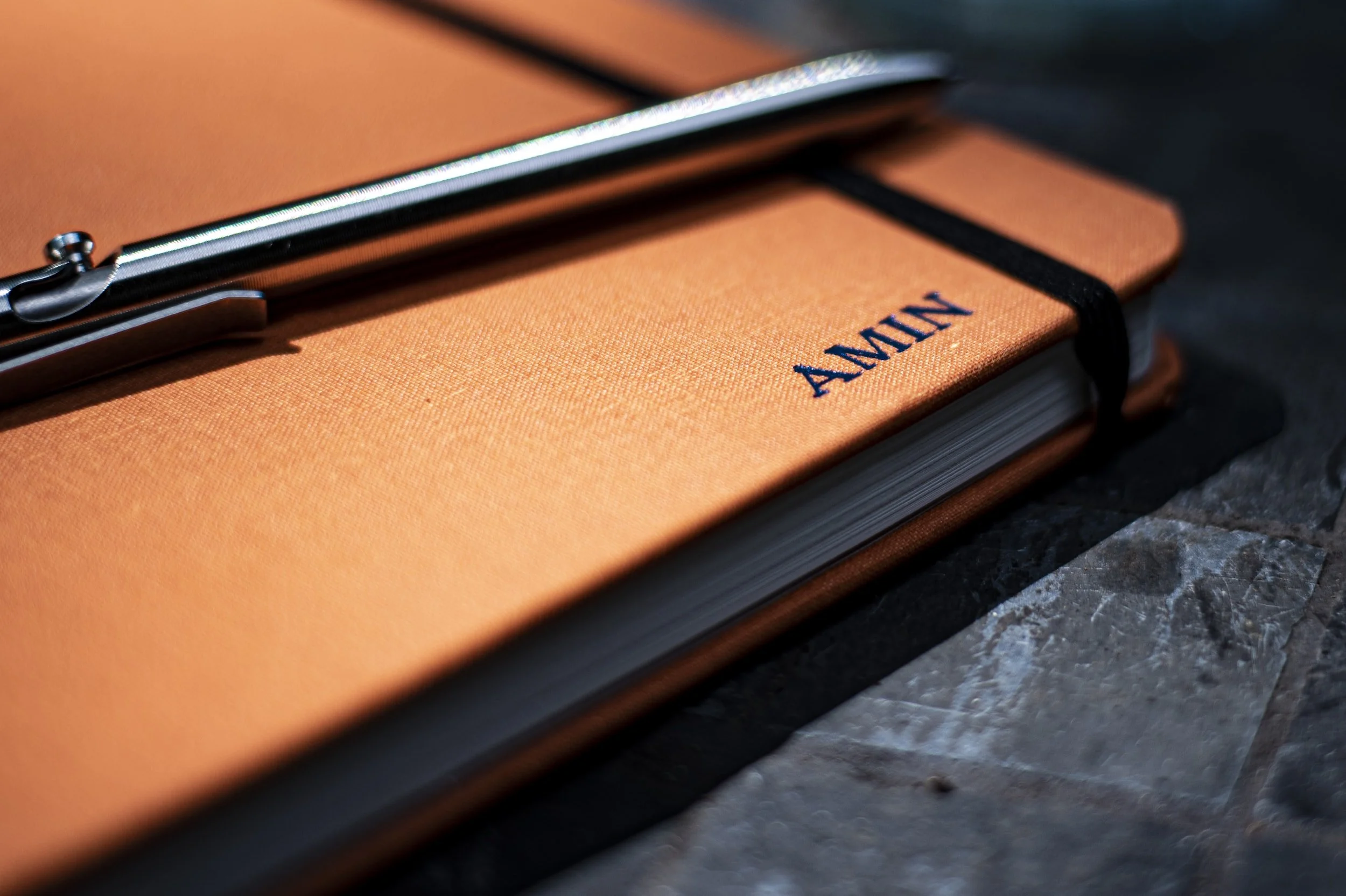 Close-up of an orange notebook with "AMIN" embossed, placed on a stone surface, with a silver pen resting on top.