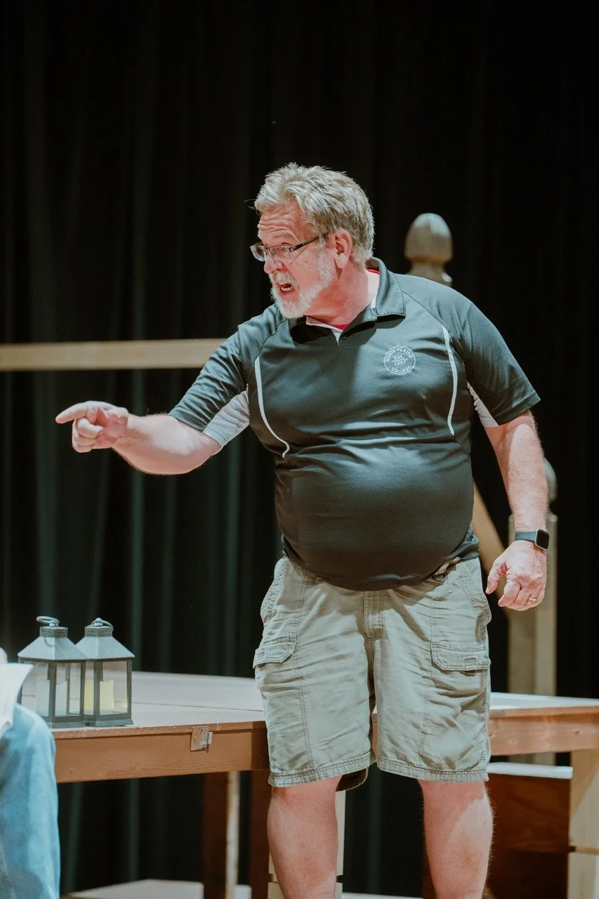 Man in a short-sleeved shirt and shorts gesturing on stage during a performance, with two lanterns on a table.