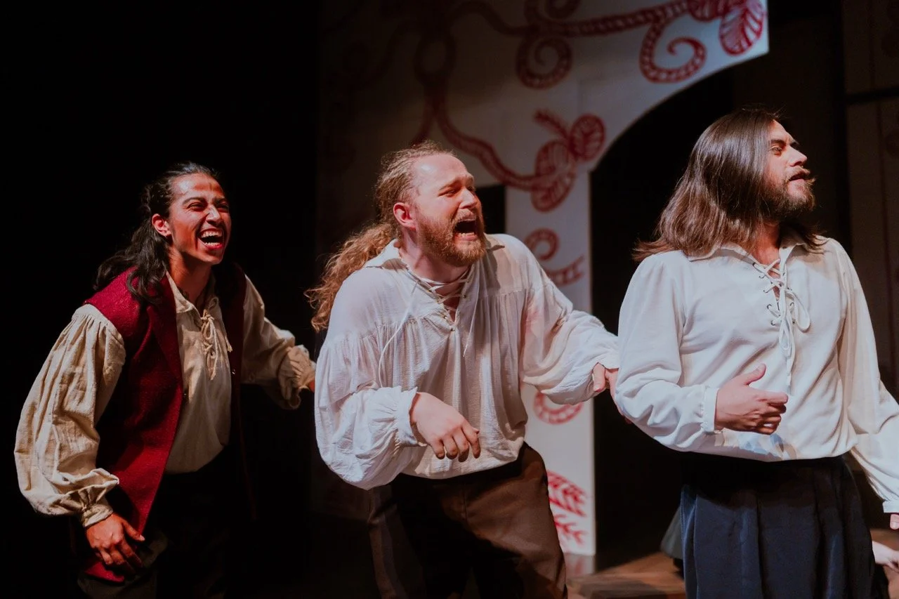 Three actors performing on stage in period costumes with expressive faces, against a backdrop with red patterns.