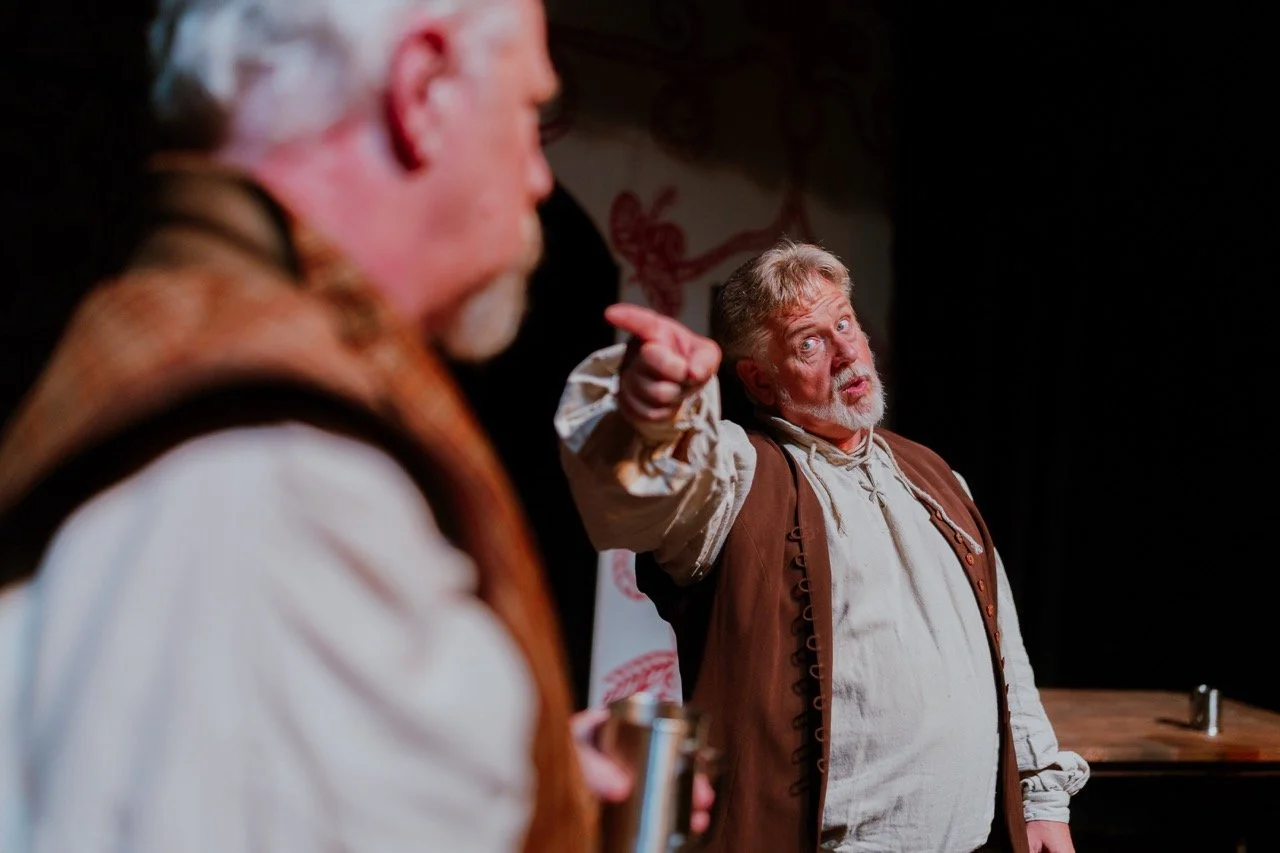 Two actors in period costumes perform on stage, one pointing and the other partially visible, holding a cup.