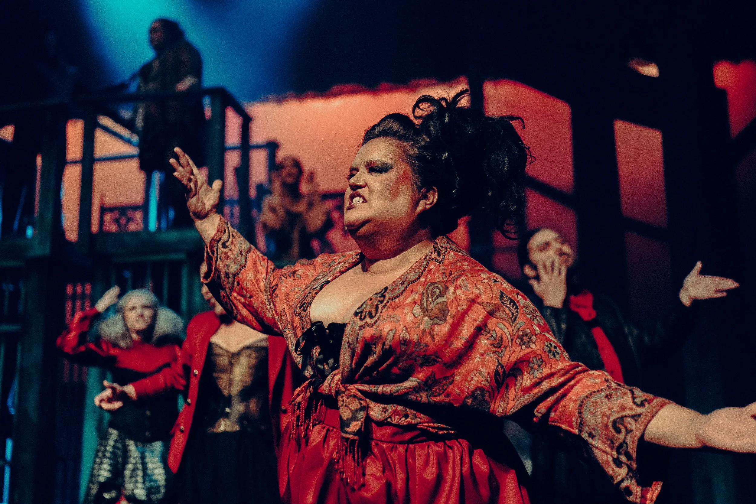 Theatrical performance scene with actors in dramatic costumes; prominent actor in foreground wearing a patterned robe, gesturing passionately, with supporting cast in background.