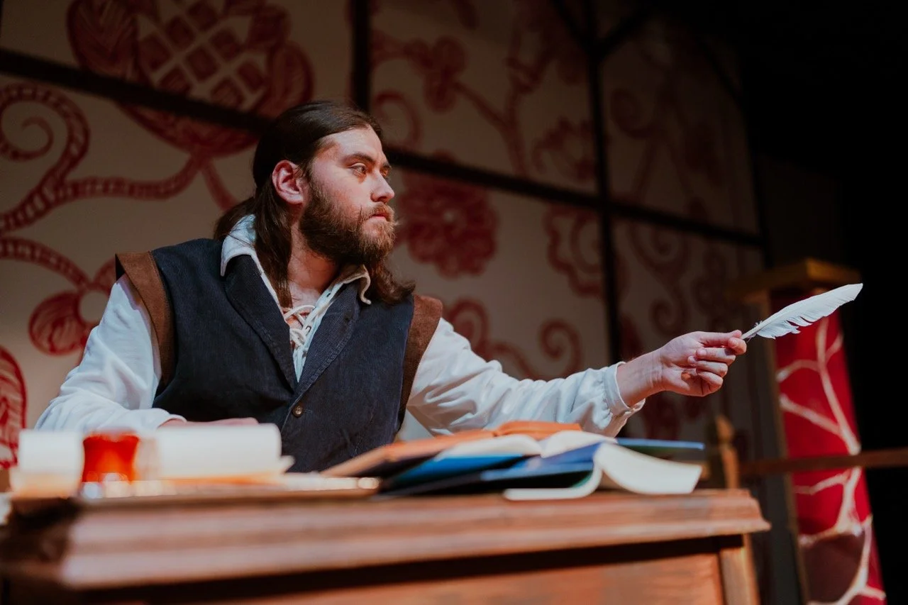 A man in historical clothing using a quill pen at a wooden desk with open books and papers.