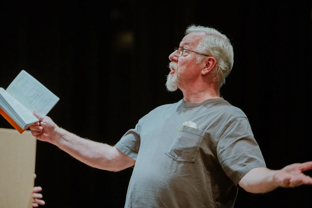 Elderly man with a gray beard reading from an open book, wearing glasses and a gray t-shirt, gesturing with one arm extended.