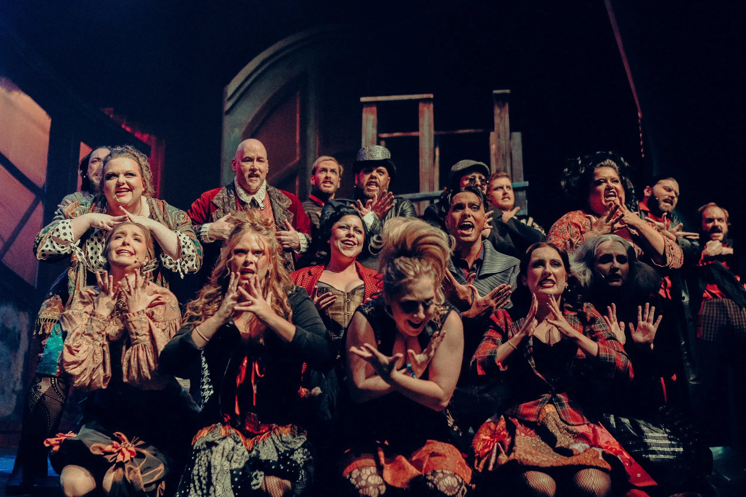 A group of theatrical performers on stage in costume, expressing dramatic emotions with their hands and faces. The scene has a vintage, Victorian-era style with diverse characters from a musical or play.