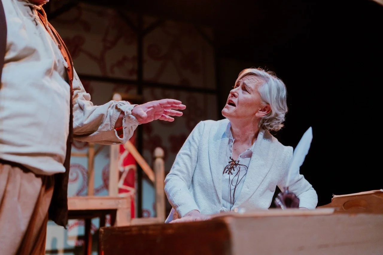Two actors performing a dramatic scene on stage, one sitting with a feather pen and the other gesturing.