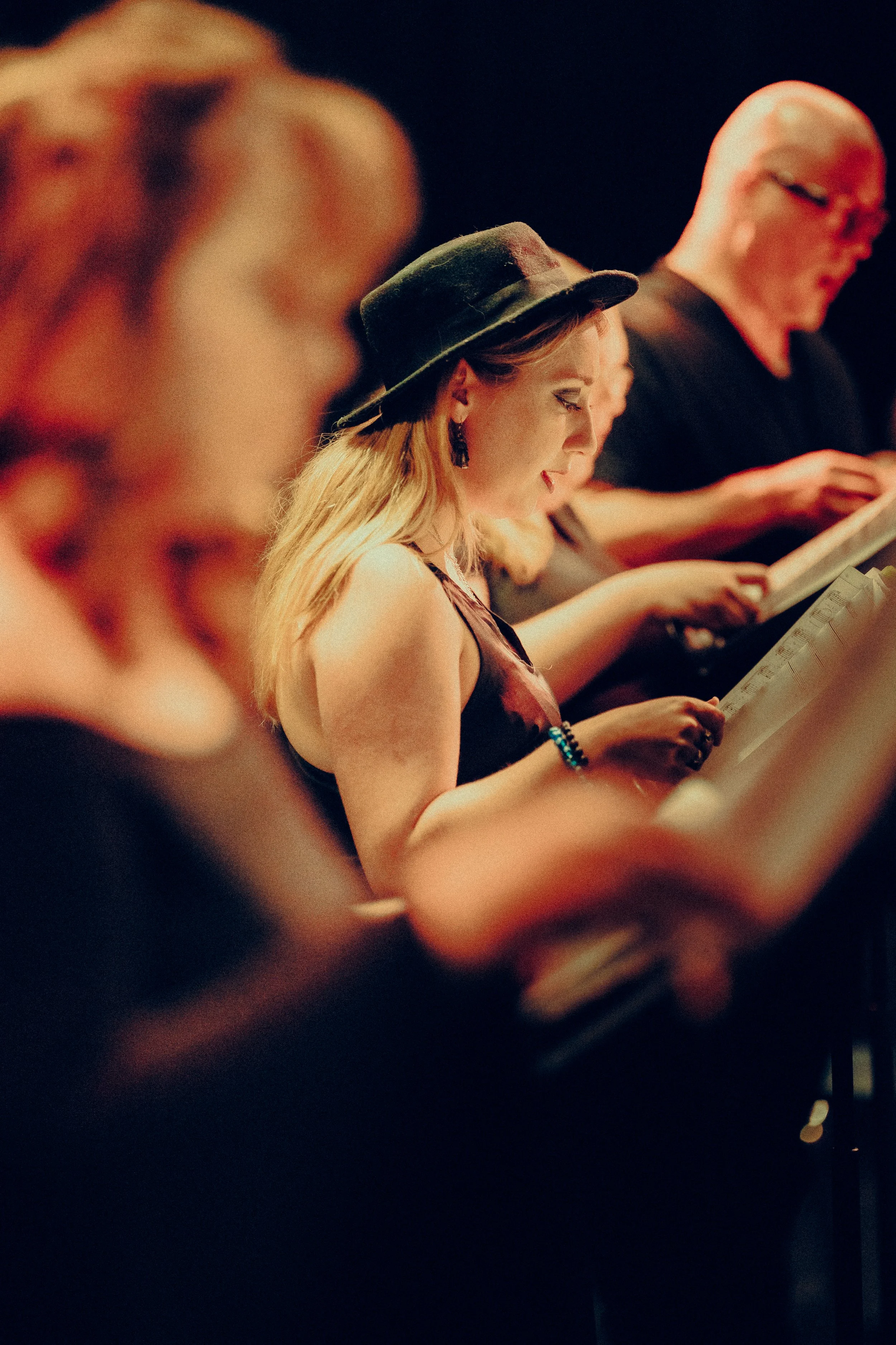 Group of people reading scripts at a theater rehearsal, woman in foreground wearing a black hat.