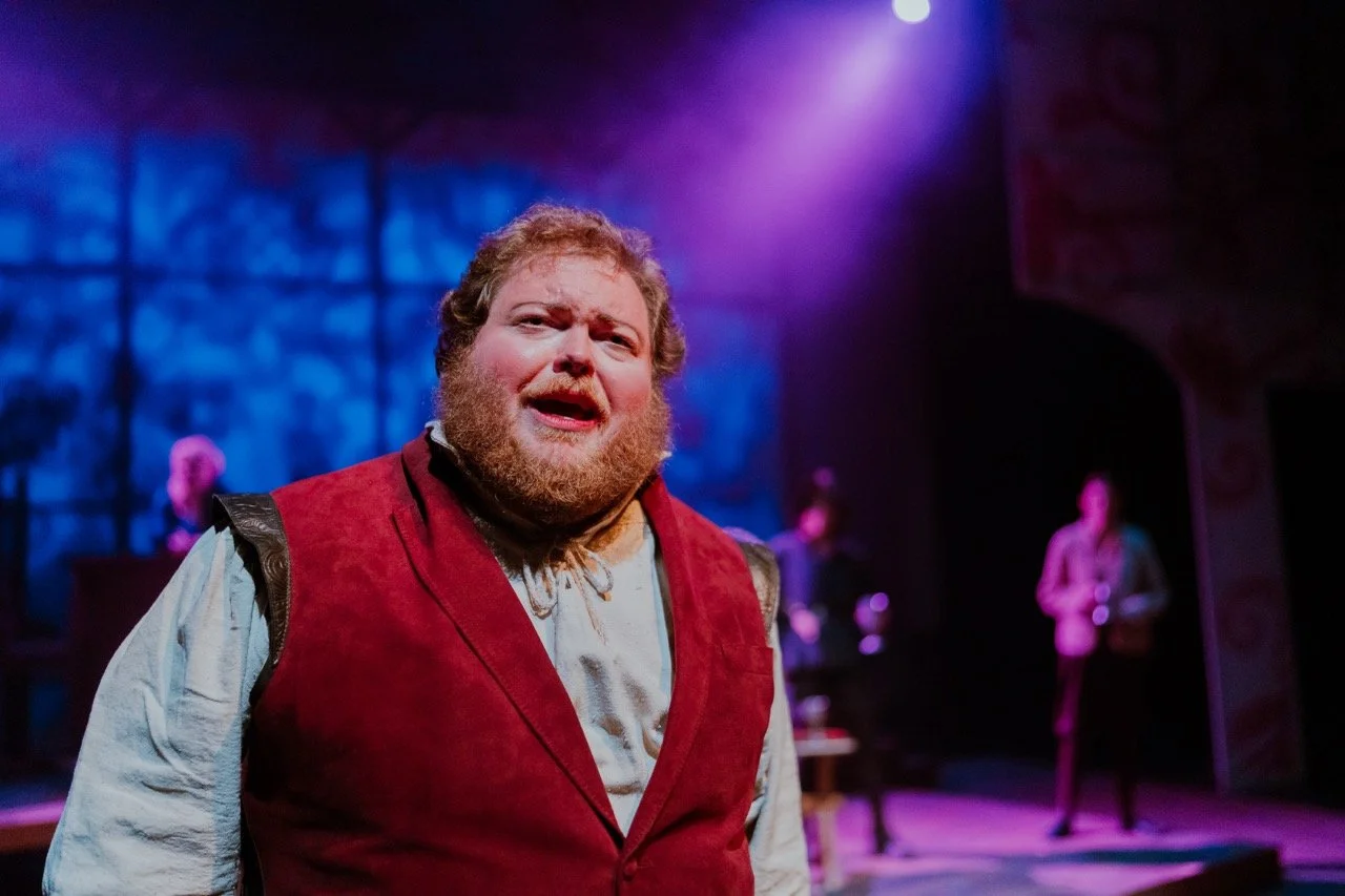 Actor on stage wearing a red vest and blue shirt with a dramatic expression under stage lights.