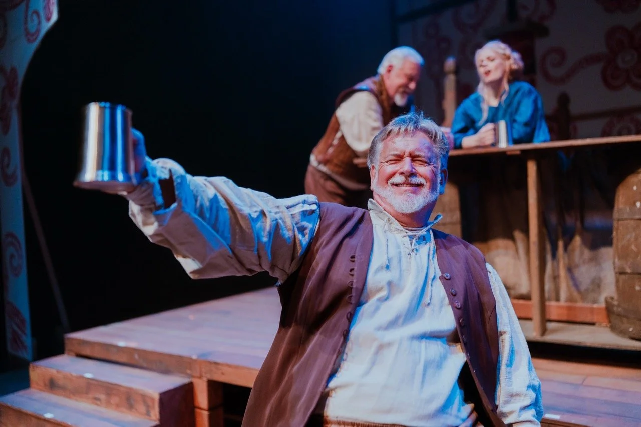 A joyful man in period costume holds a mug high in celebration on stage with two other performers in the background.