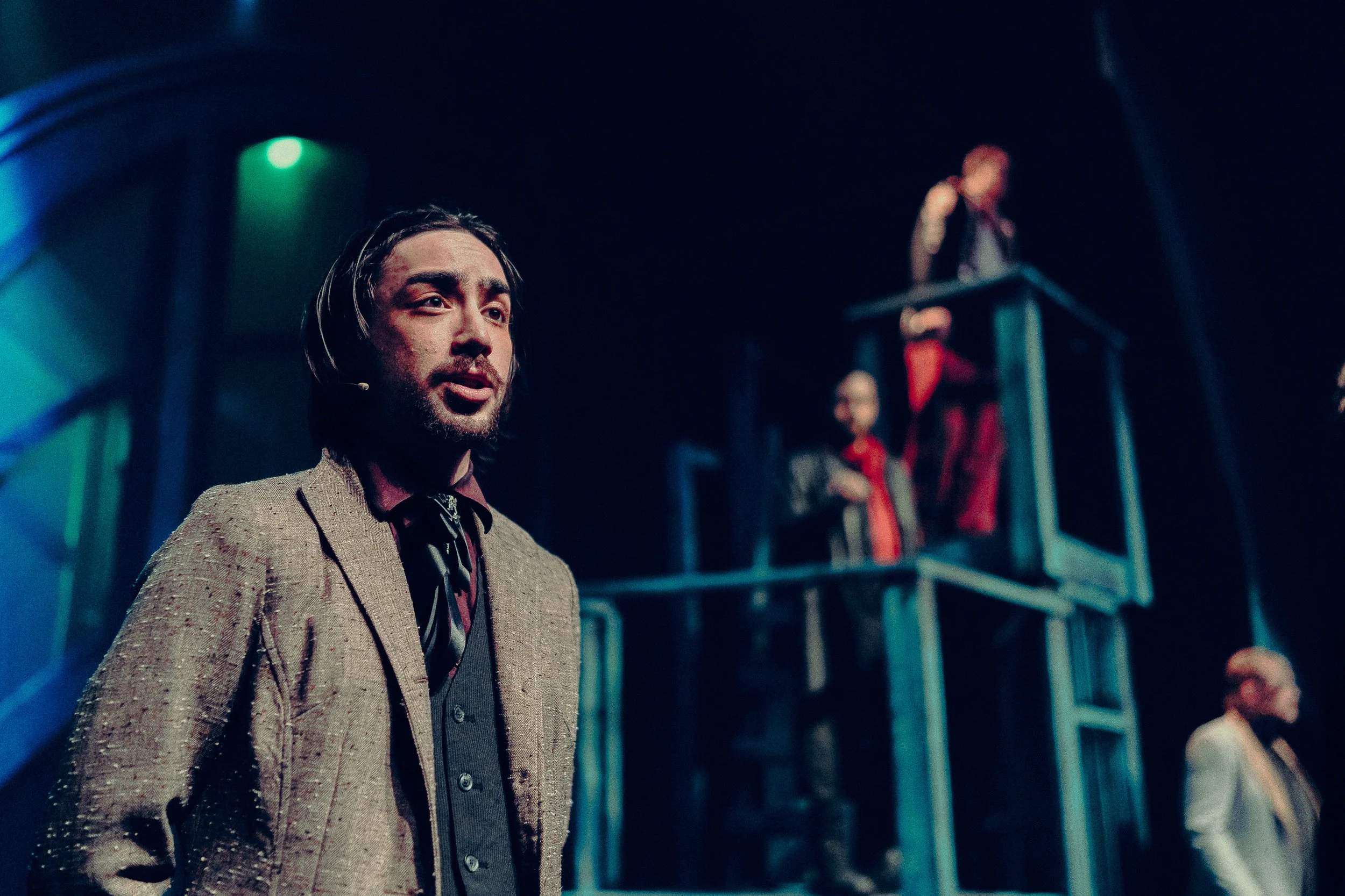 A man in a suit performing on stage with other actors in a theatrical play. The background shows stage lighting and a multi-level set structure.