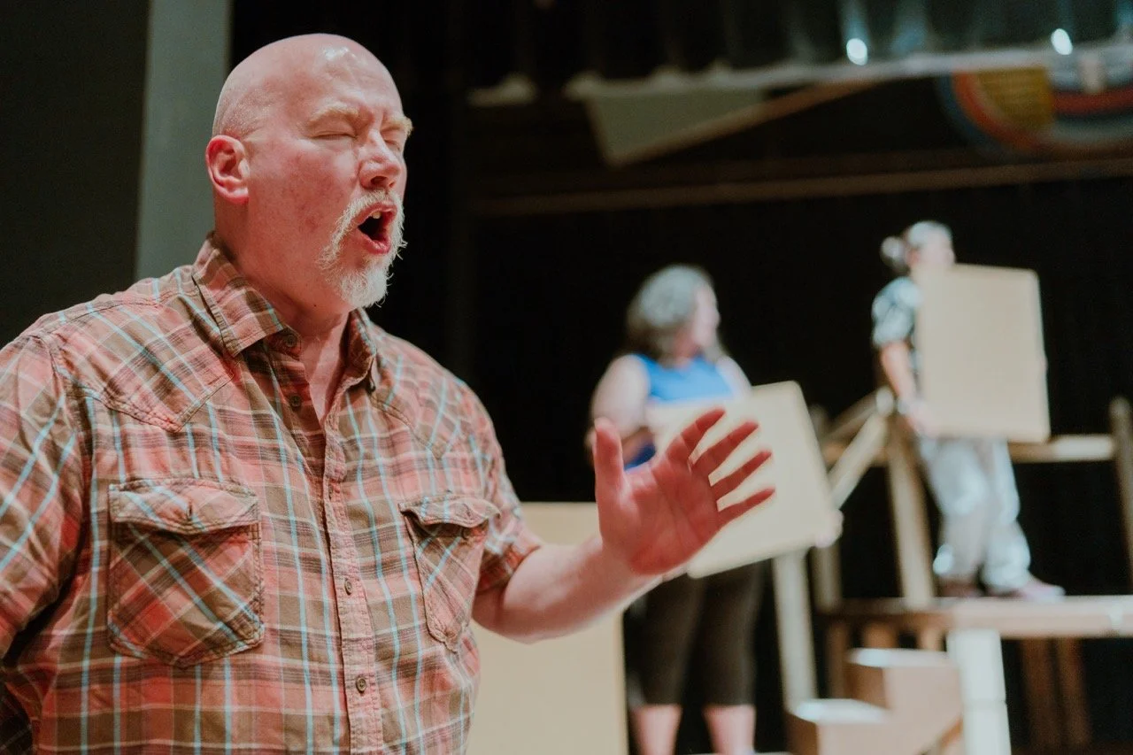 Man in plaid shirt singing or acting passionately on stage with two blurred people holding props in the background.