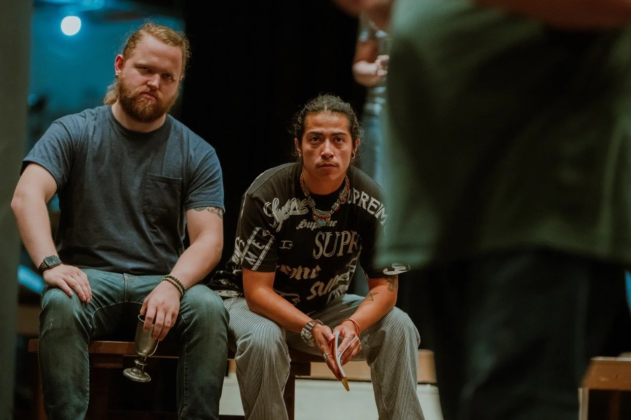 Two seated men, one with a beard holding a glass, the other wearing a patterned shirt, both looking intently forward, in an indoor setting.