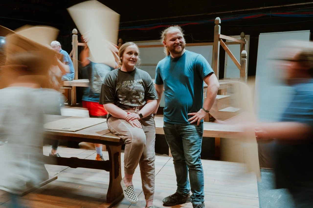 Two people on stage with backdrop; others moving around carrying props; wooden structure background.