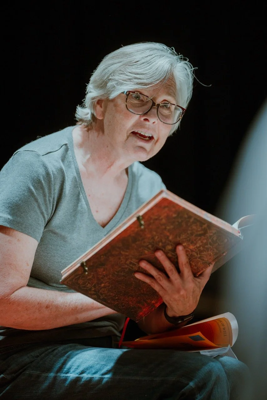 Elderly person with gray hair and glasses reading a book in a dark setting, wearing a gray shirt.