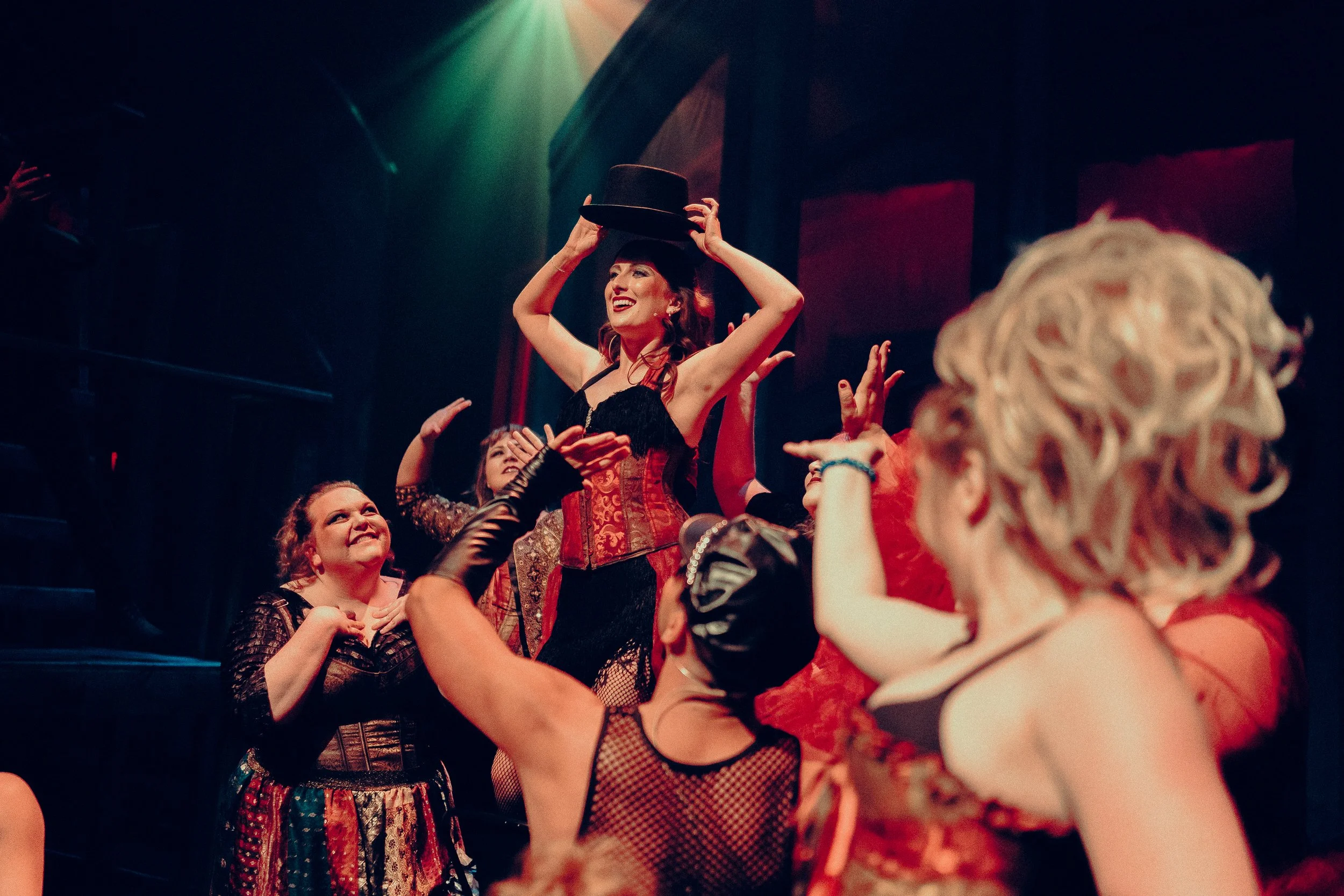 Group of performers on stage in colorful costumes with one holding a hat, in a theatrical setting.
