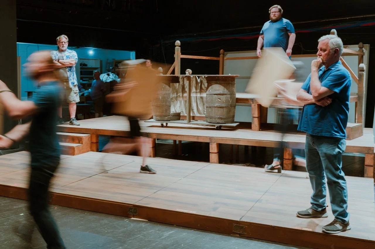 A theater stage with people rehearsing, including a man thinking and another holding a clipboard, blurred movement of others in the foreground, and wooden barrels on the stage.