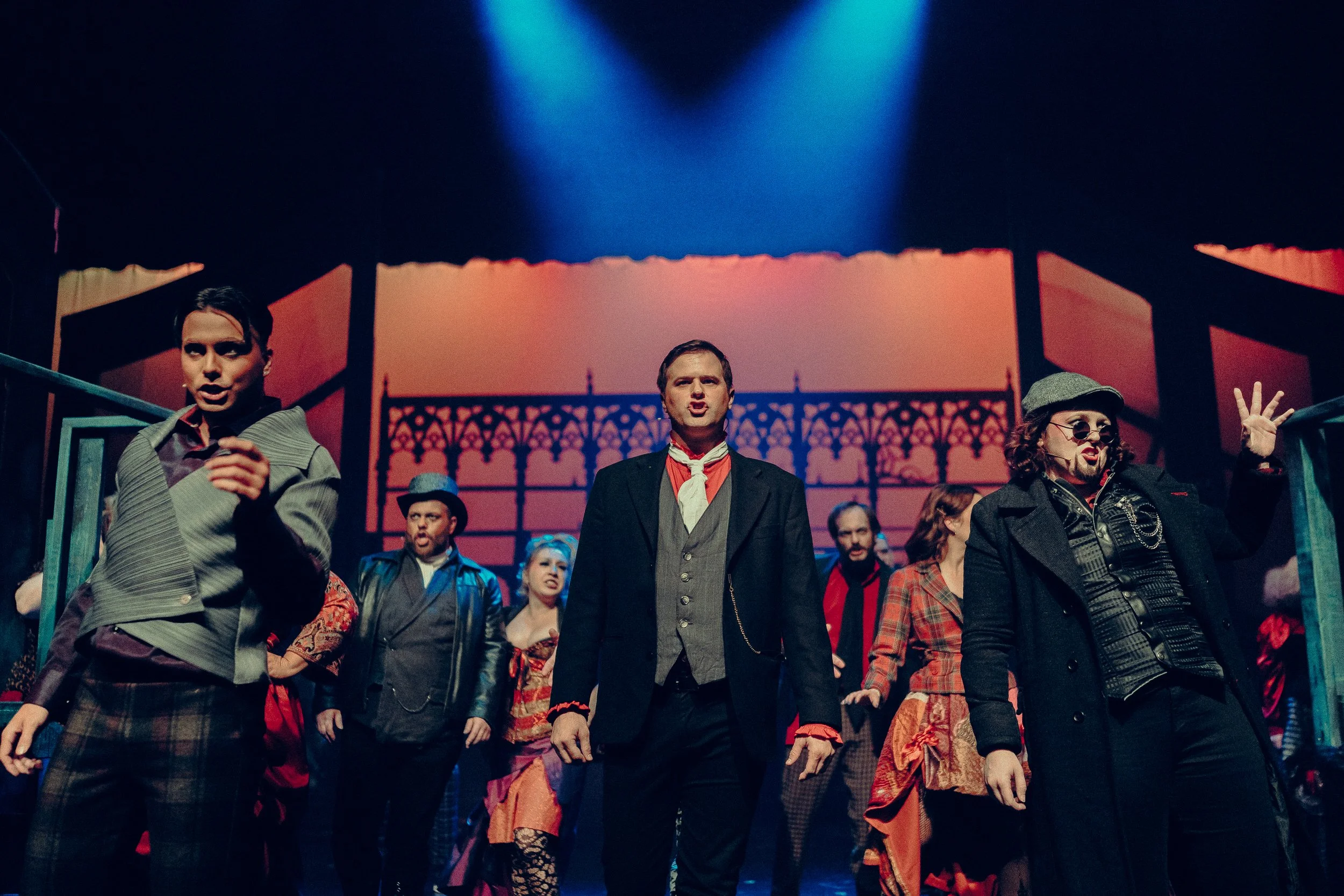 A group of performers in theatrical costumes on stage, with dramatic lighting and a decorative backdrop. The central figure wears a suit with a red scarf, flanked by others in various period-style garments and accessories. Blue and orange lights high