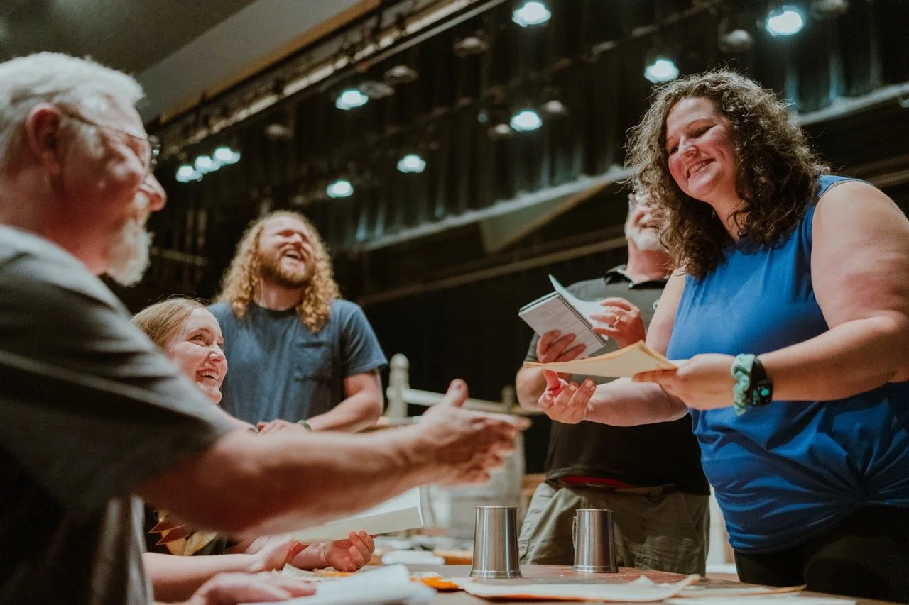 Group of people laughing and interacting in a theater setting with scripts in hand