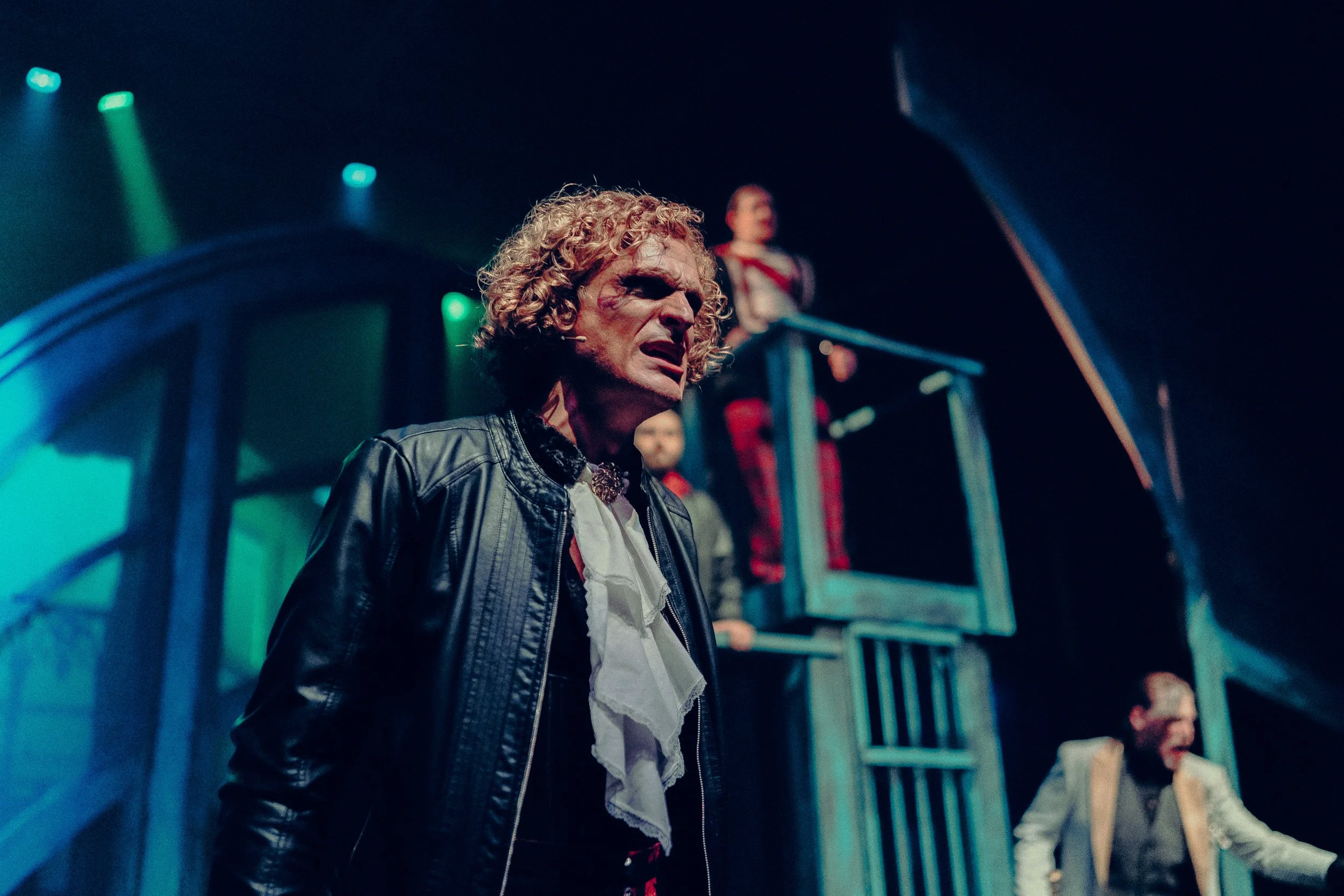 A theatrical performance with a man wearing a leather jacket and white ruffled shirt on stage, expressive lighting, two other performers in the background, one elevated.