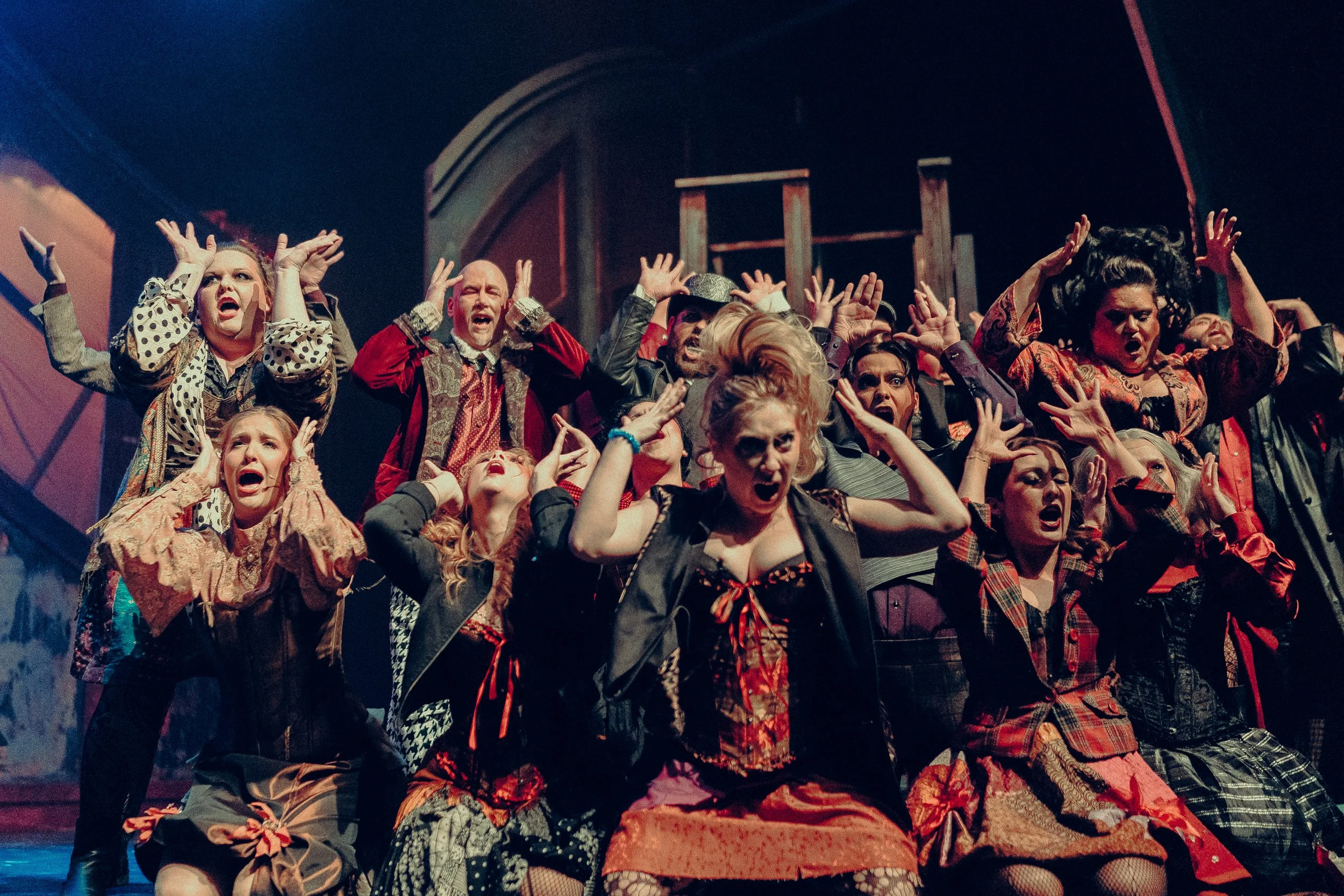 Theatrical performance with a diverse group of actors in expressive costumes and dramatic poses, performing on stage with a dark, dramatic backdrop.