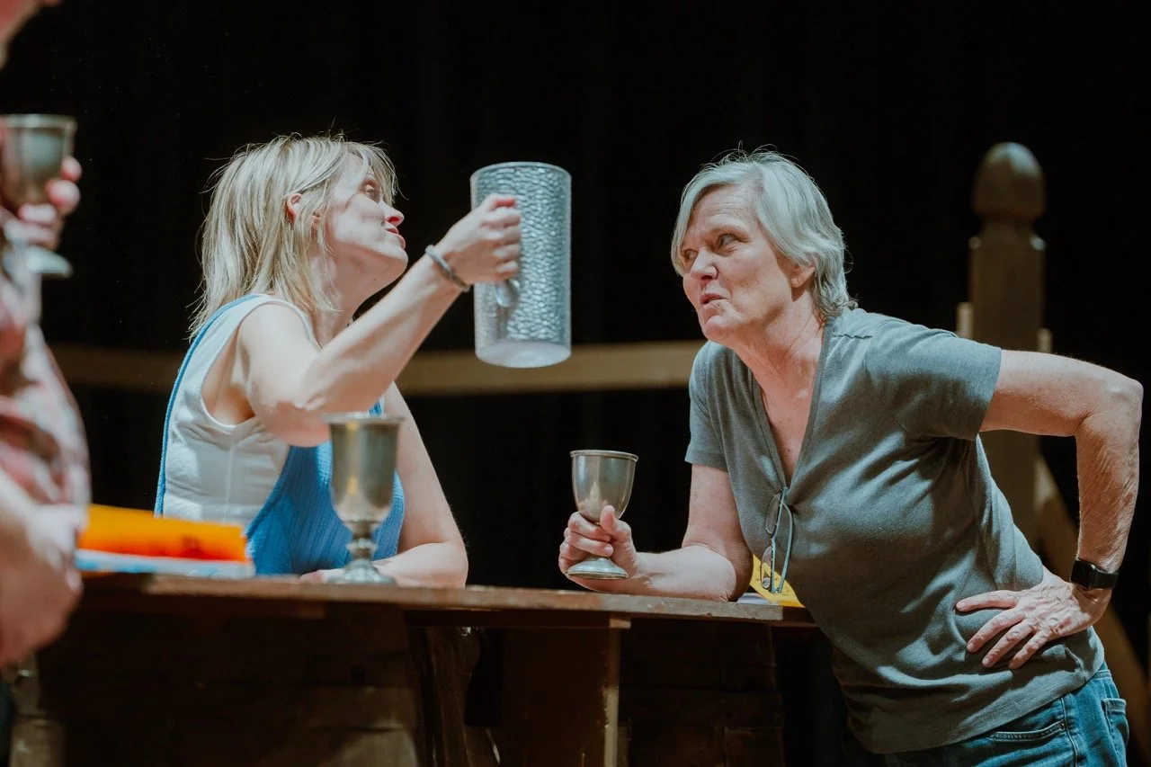 Two women engaging in conversation at a table, holding metal goblets, with one of them raising a textured pitcher.