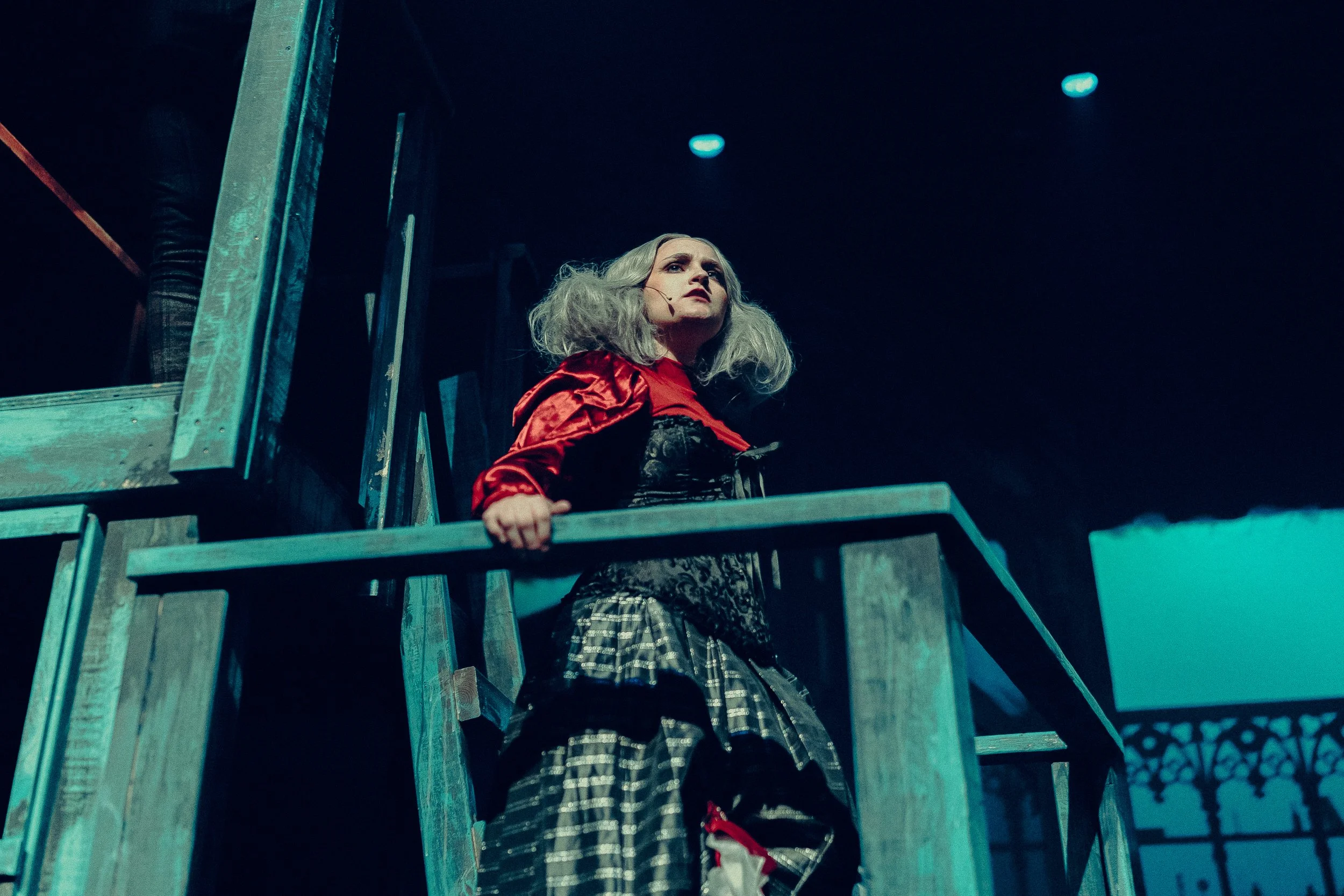 A woman in theatrical costume with silver hair and a red and black outfit stands on a blue wooden structure, possibly part of a stage set.