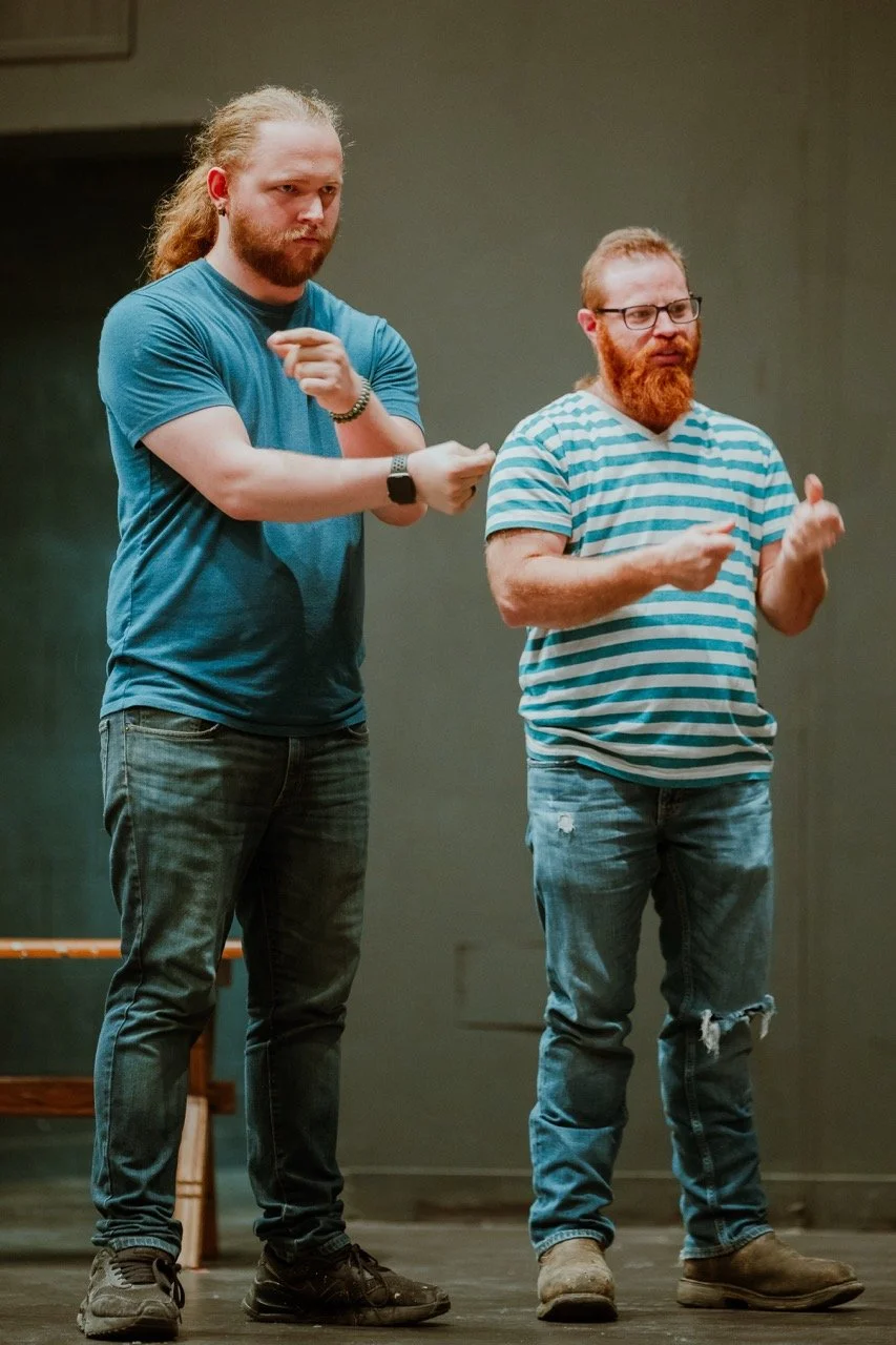Two men in casual clothing, one wearing a plain blue shirt and the other a blue striped shirt, standing and gesturing with their hands.