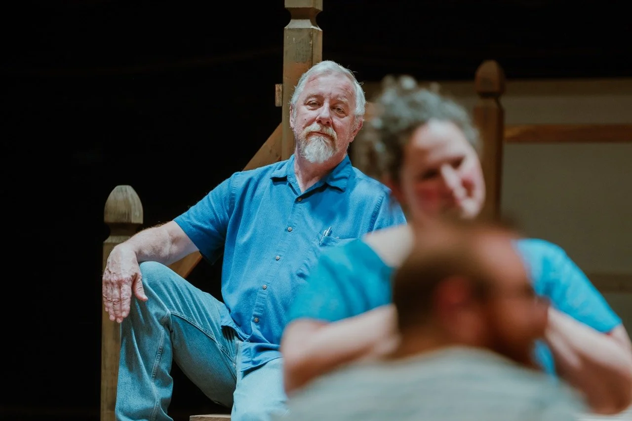 Elderly man sitting on a wooden staircase, wearing a blue shirt and jeans, with other people blurred in the foreground.