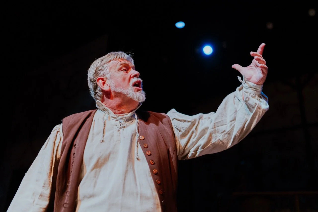 Actor performing on stage under dramatic lighting, wearing a historical costume with a vest and long-sleeved shirt, gesturing expressively.