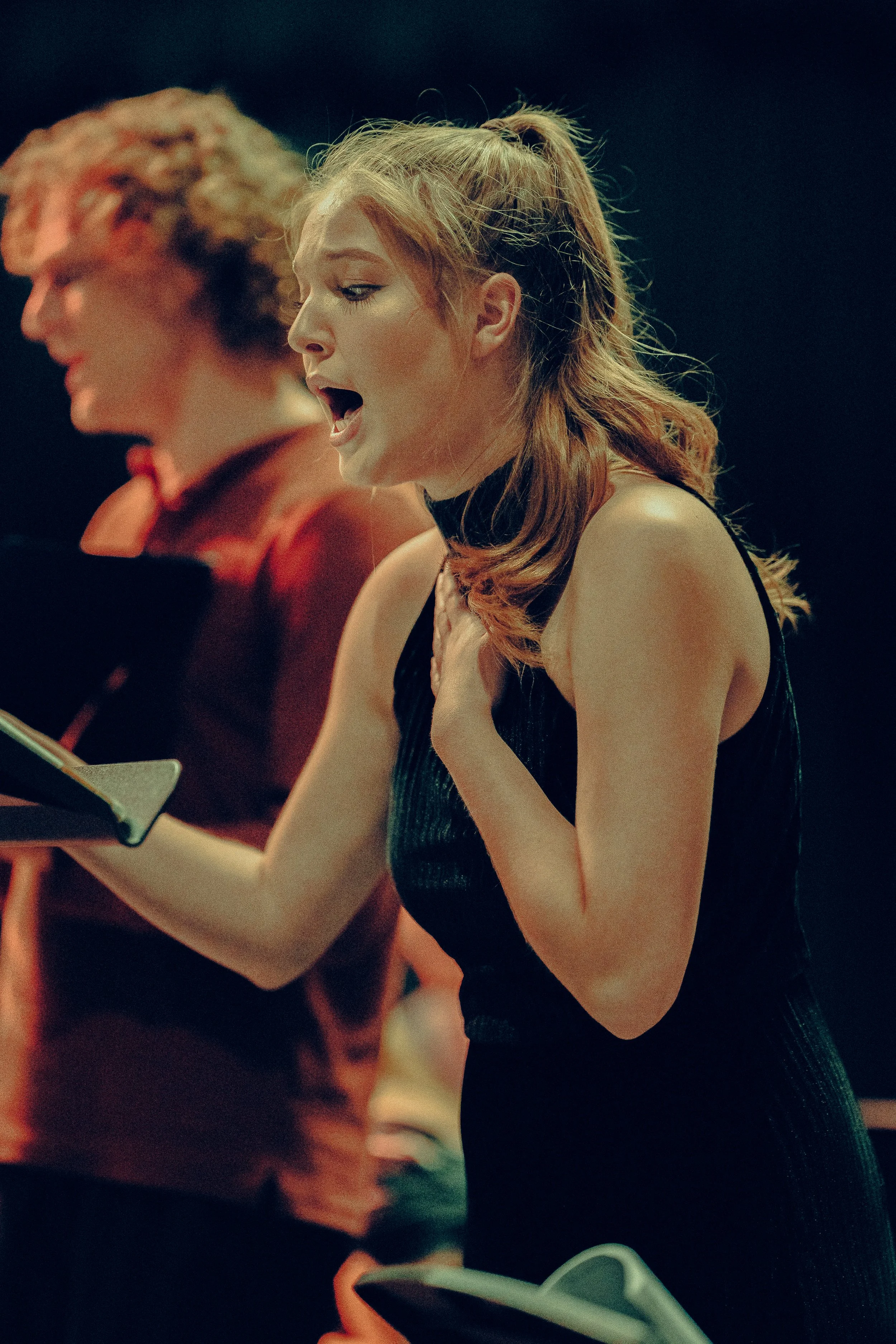 A young woman singing passionately with sheet music in hand during a performance. Another person is visible in the background.