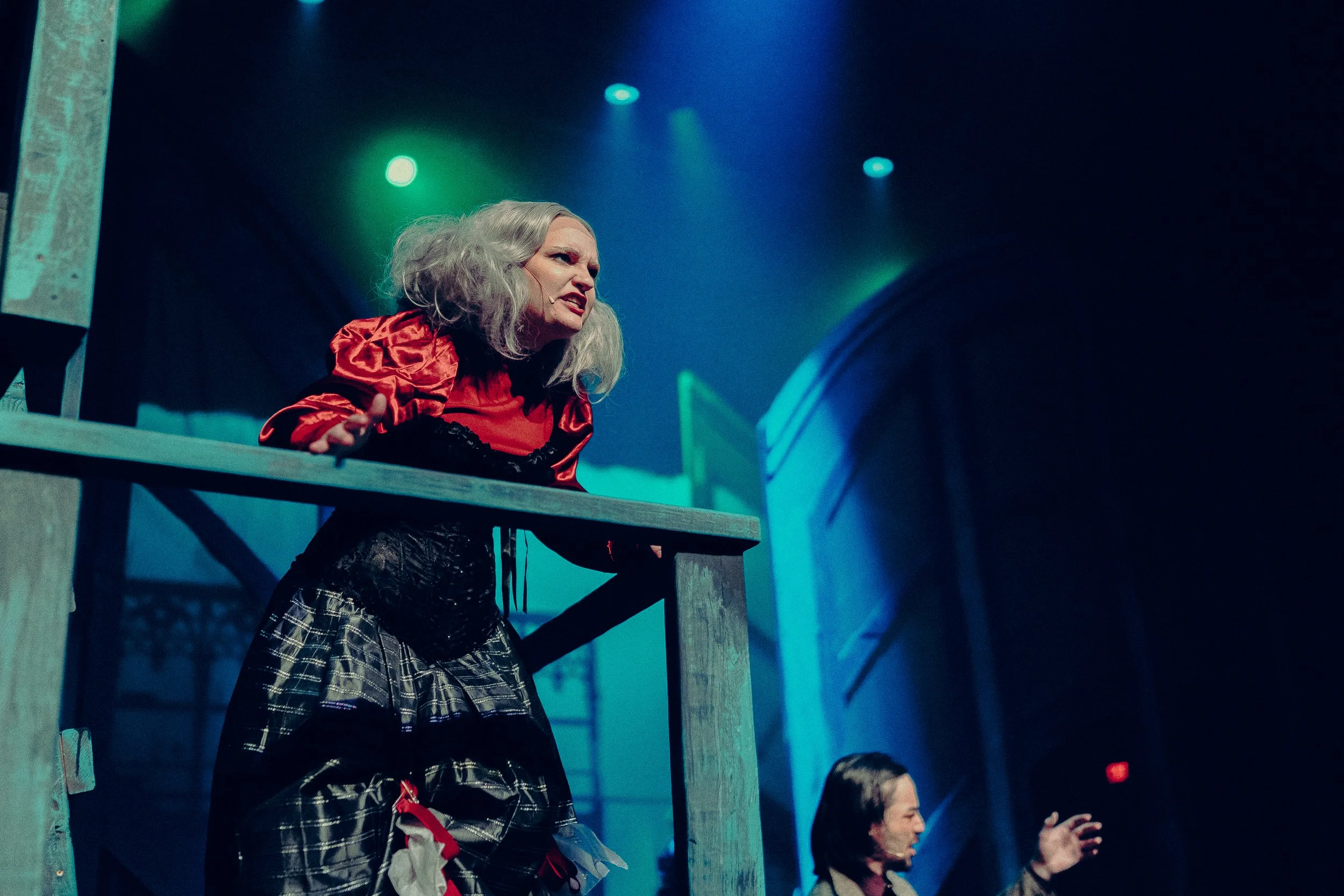 Theatrical scene with a performer in a red and black costume acting on stage, under colorful spotlights, with a dramatic expression.