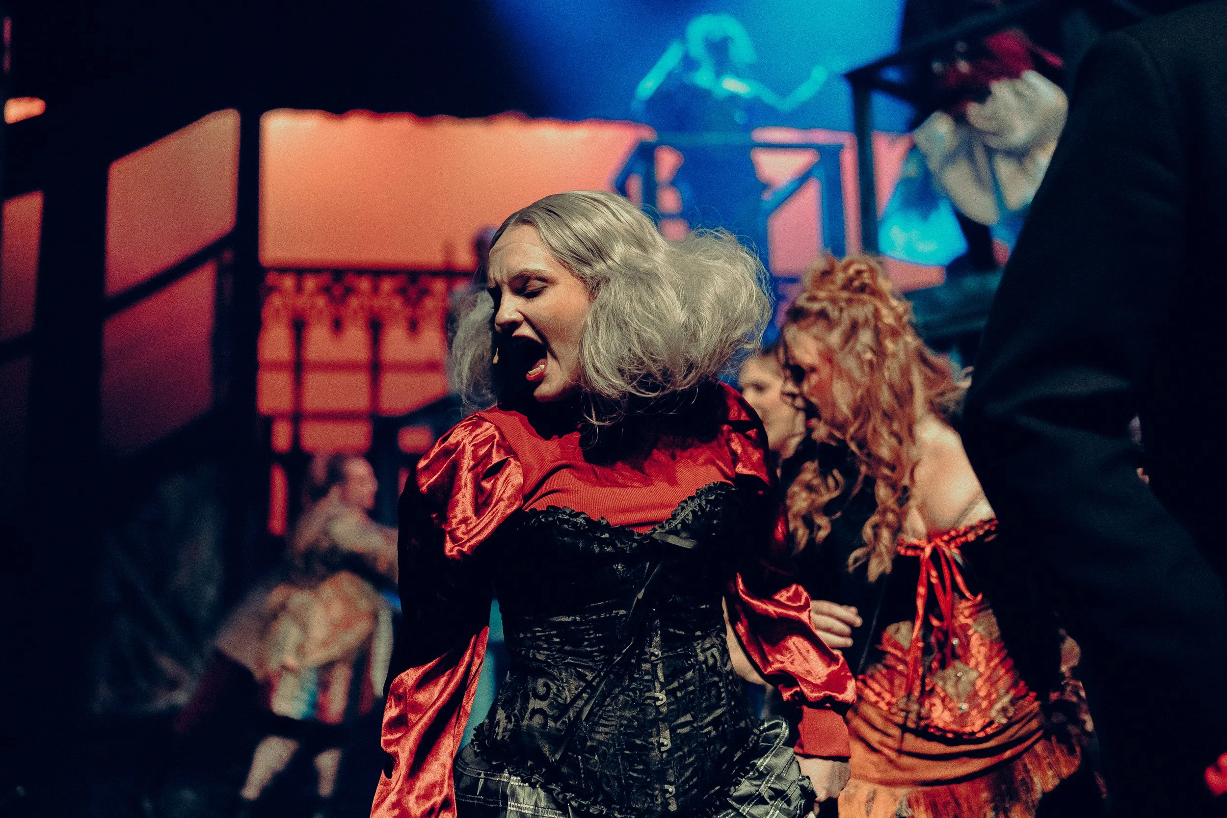 Woman in theatrical costume with gray wig performing on stage with dramatic lighting.