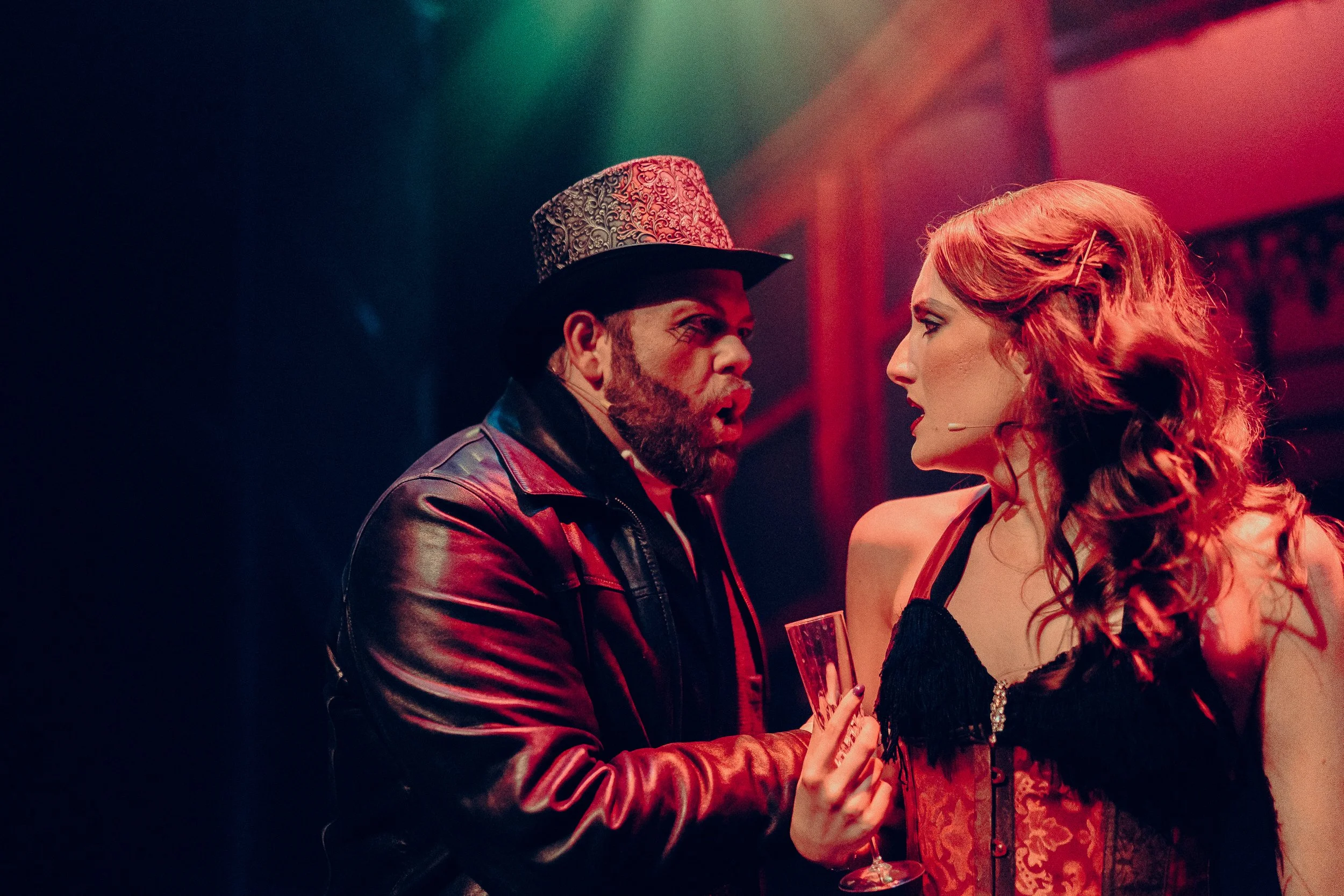 Two actors performing on stage, one wearing a decorated top hat and leather jacket, the other in a corset holding a champagne flute, under dramatic lighting.