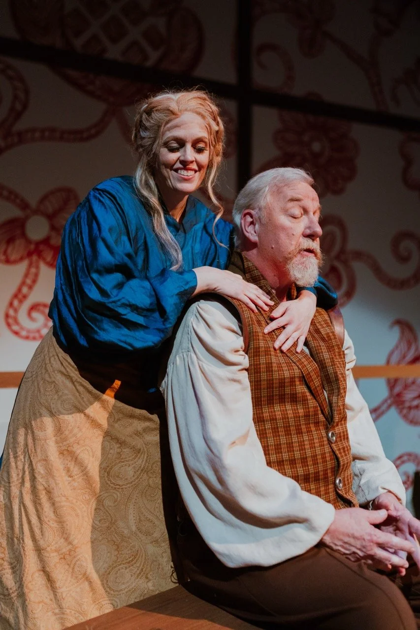 A woman in a blue blouse and tan skirt stands behind a seated elderly man with a beard, wearing a plaid vest. She has her hands on his shoulders. They appear to be in a theatrical setting with patterned walls in the background.