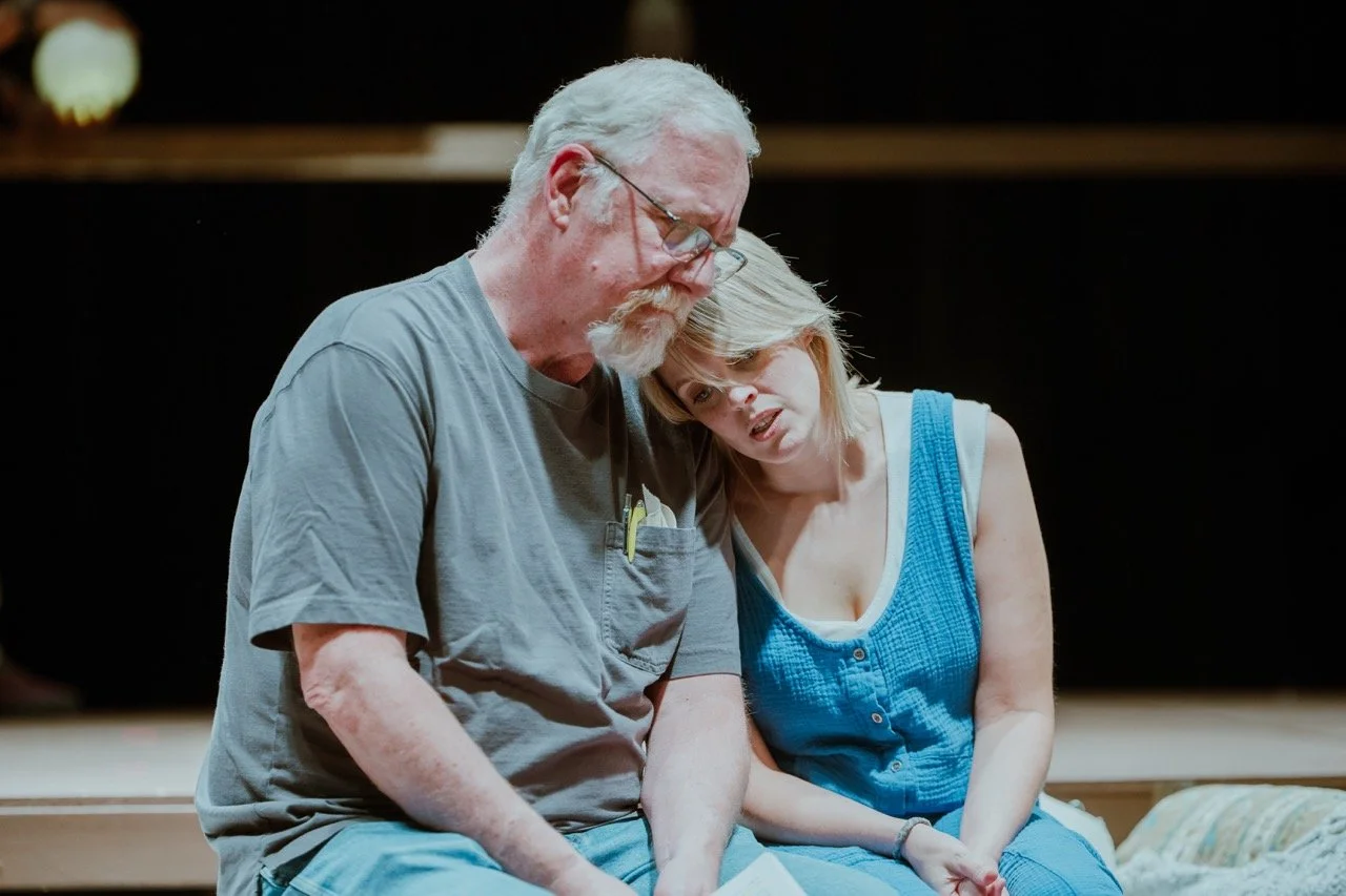 A man and a woman sitting closely on a bench, with the woman resting her head on the man's shoulder, conveying a comforting and emotional moment.