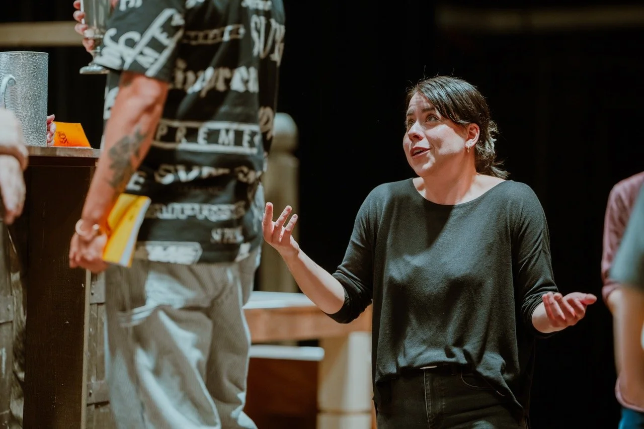A woman gesturing with hands up, looking confused, during a conversation with a man holding a book. The setting appears to be indoors with dim lighting.