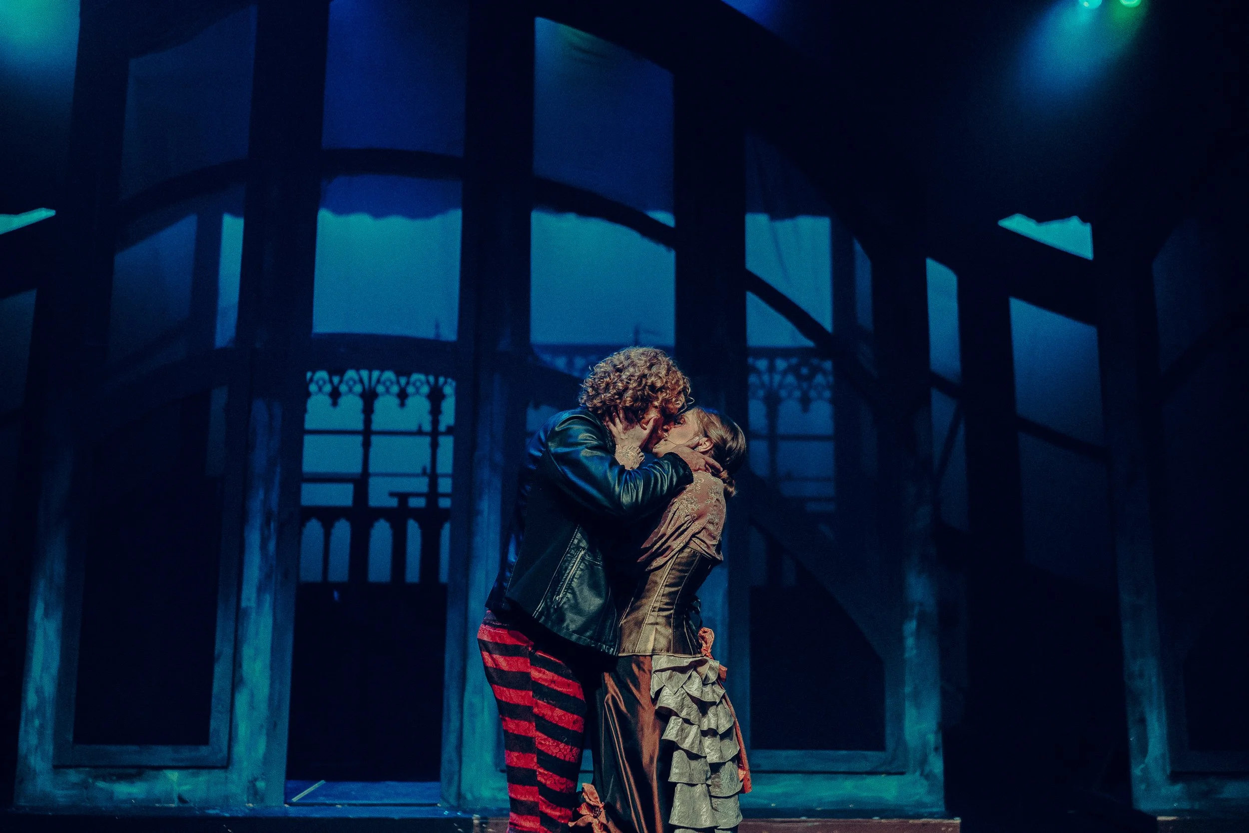 Two actors kissing on stage during a theatrical performance, with dramatic lighting and a scenic backdrop.