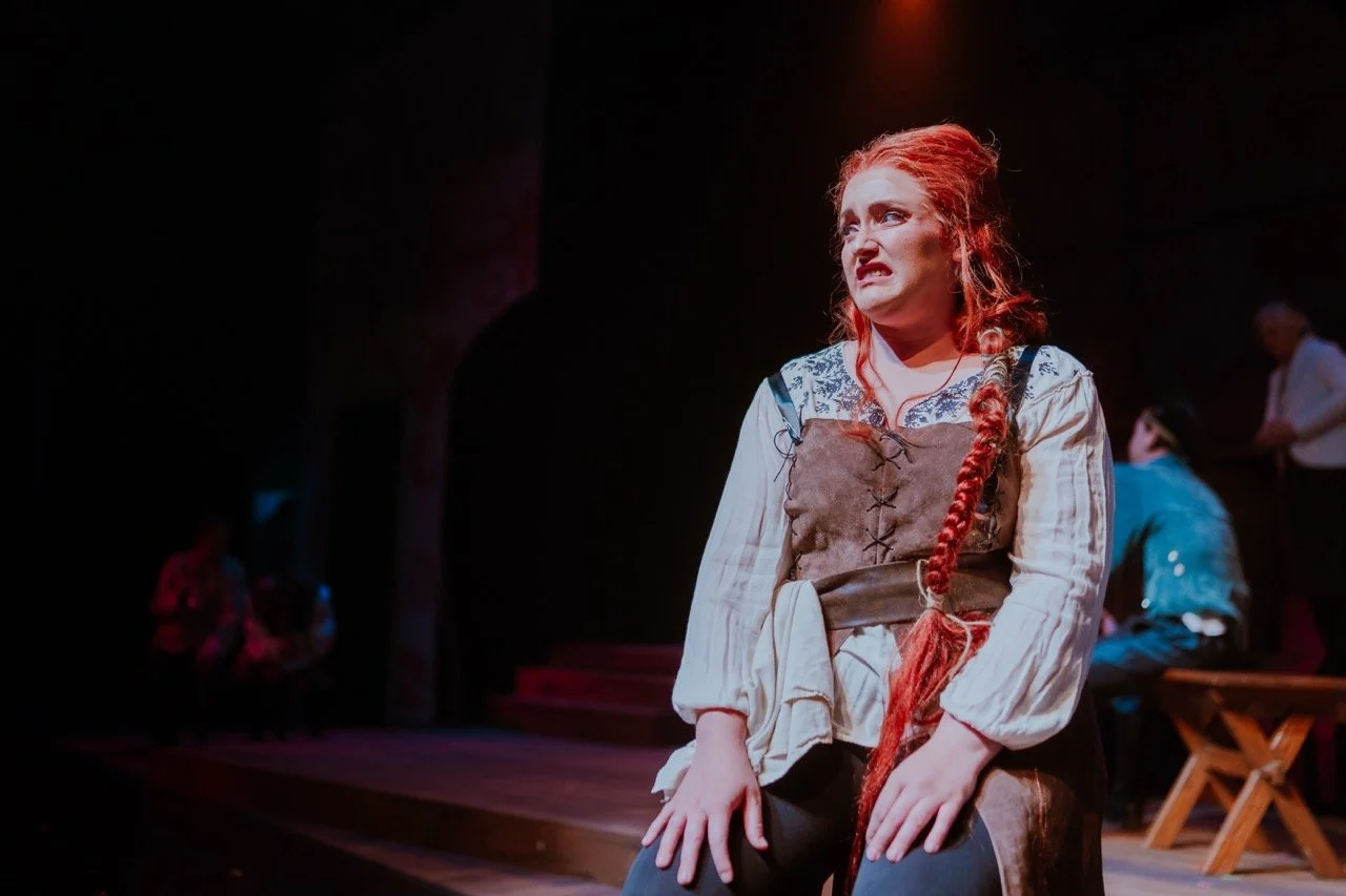 A theater actress with long red hair and a braided hairstyle performing on stage, wearing a medieval-style costume. The background is dark with a spotlight shining on her, and other performers are blurred in the distance.