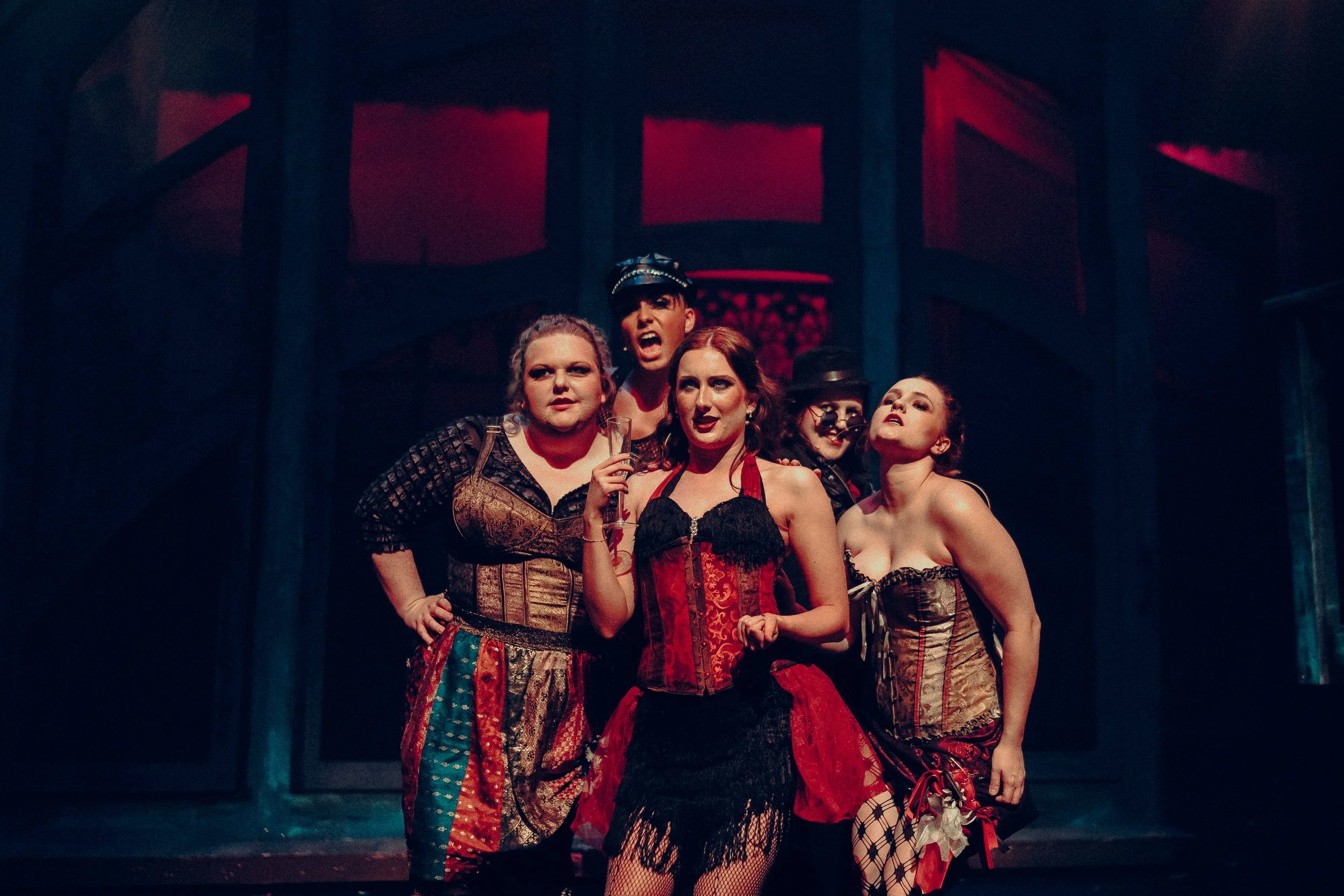 Group of performers in theatrical costumes on stage with a moody, dark background.