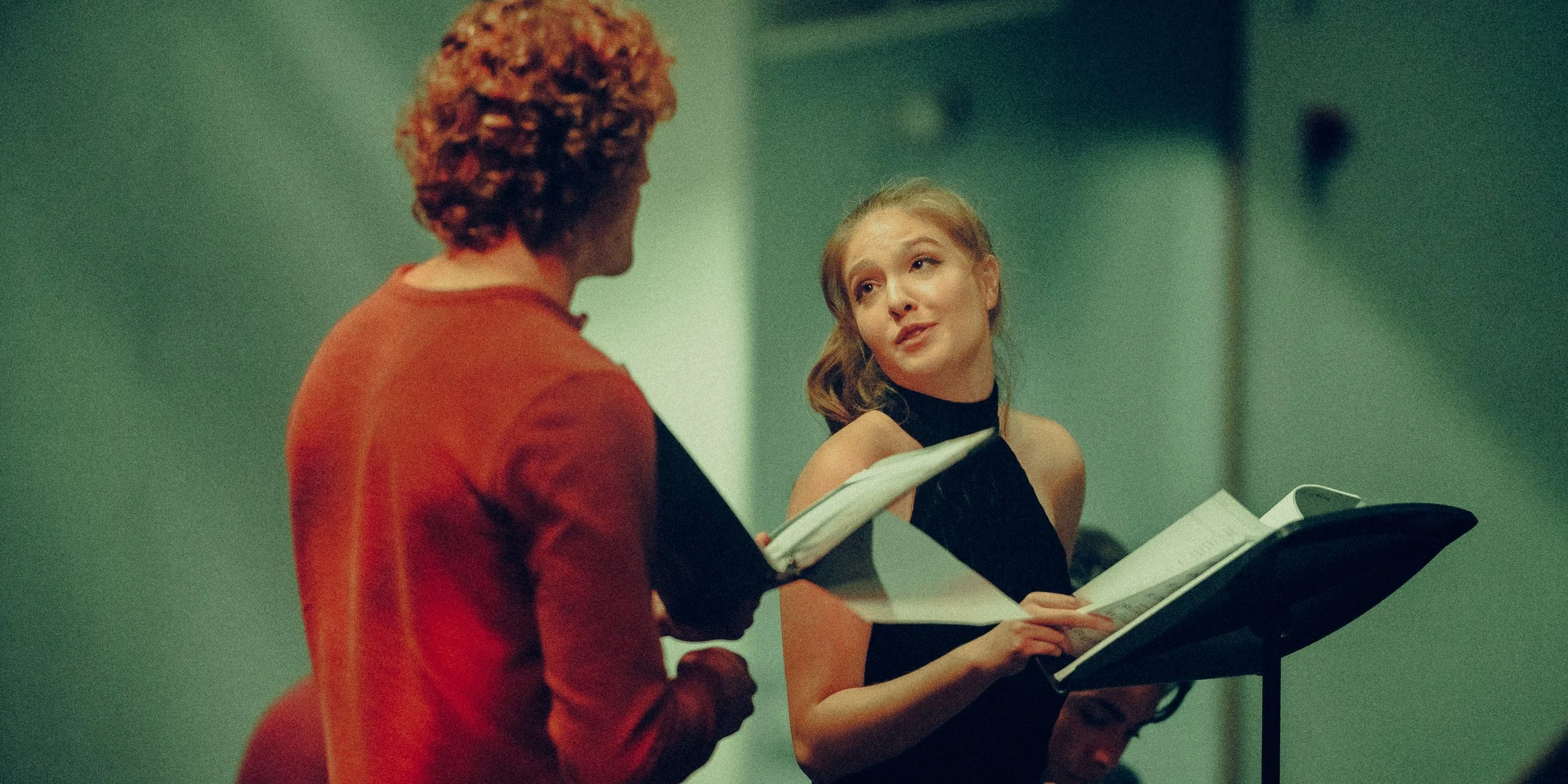 Two people with scripts, likely rehearsing a scene, one person speaking intently, the other listening.