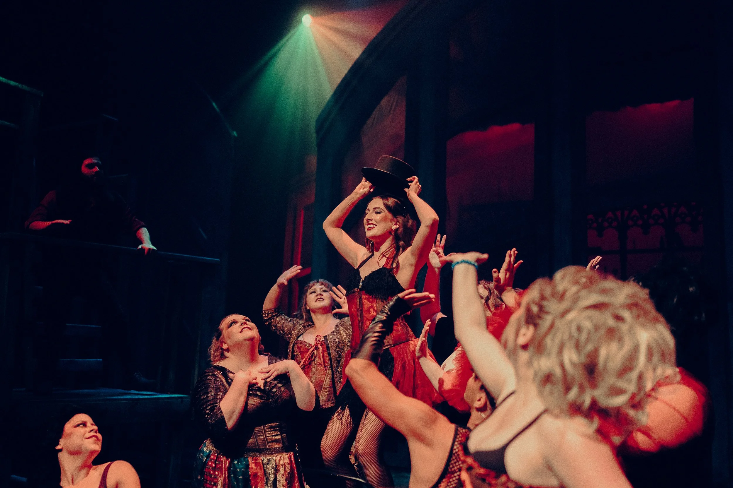 Theatrical performance with performers in colorful costumes and dramatic lighting, centered around a woman holding a top hat.
