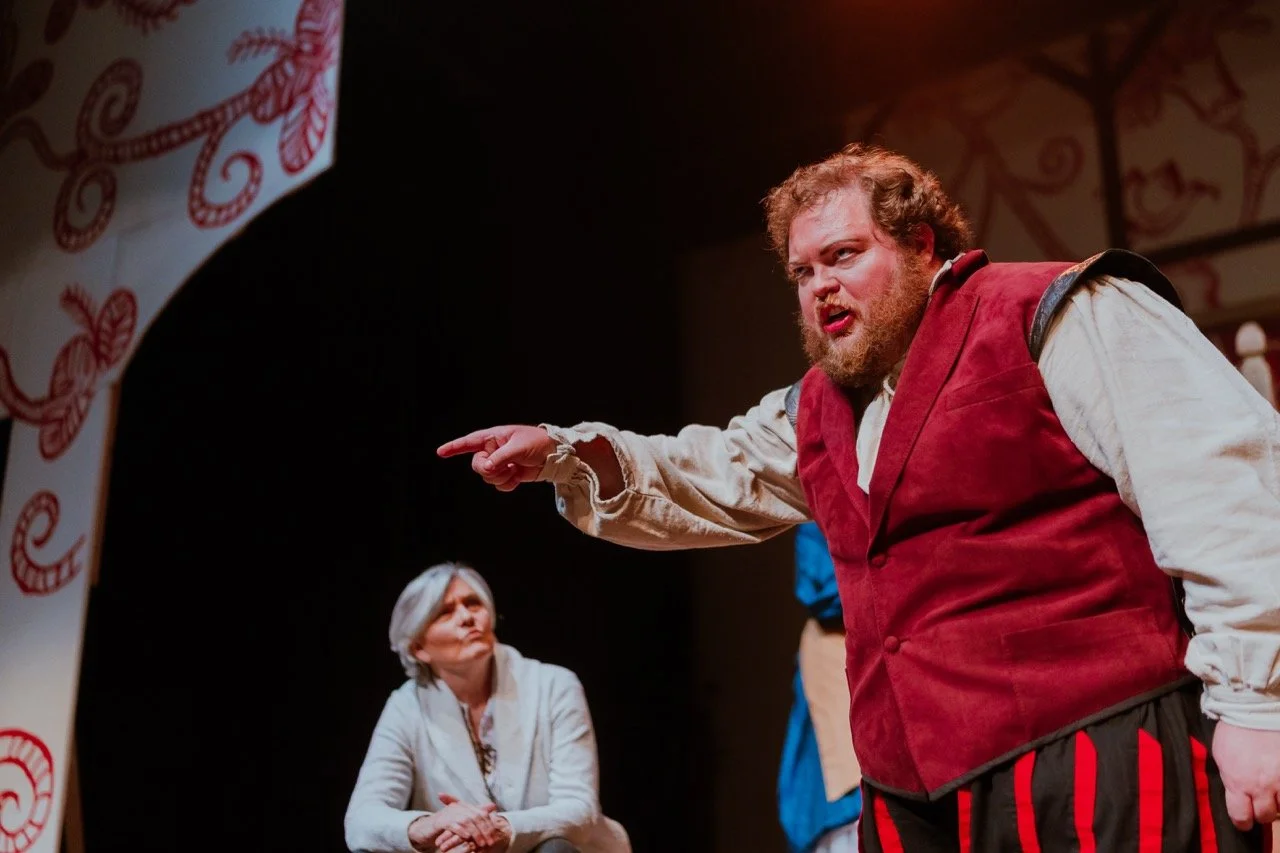 Actor in period costume performing on stage, pointing dramatically, with a seated woman observing in the background.