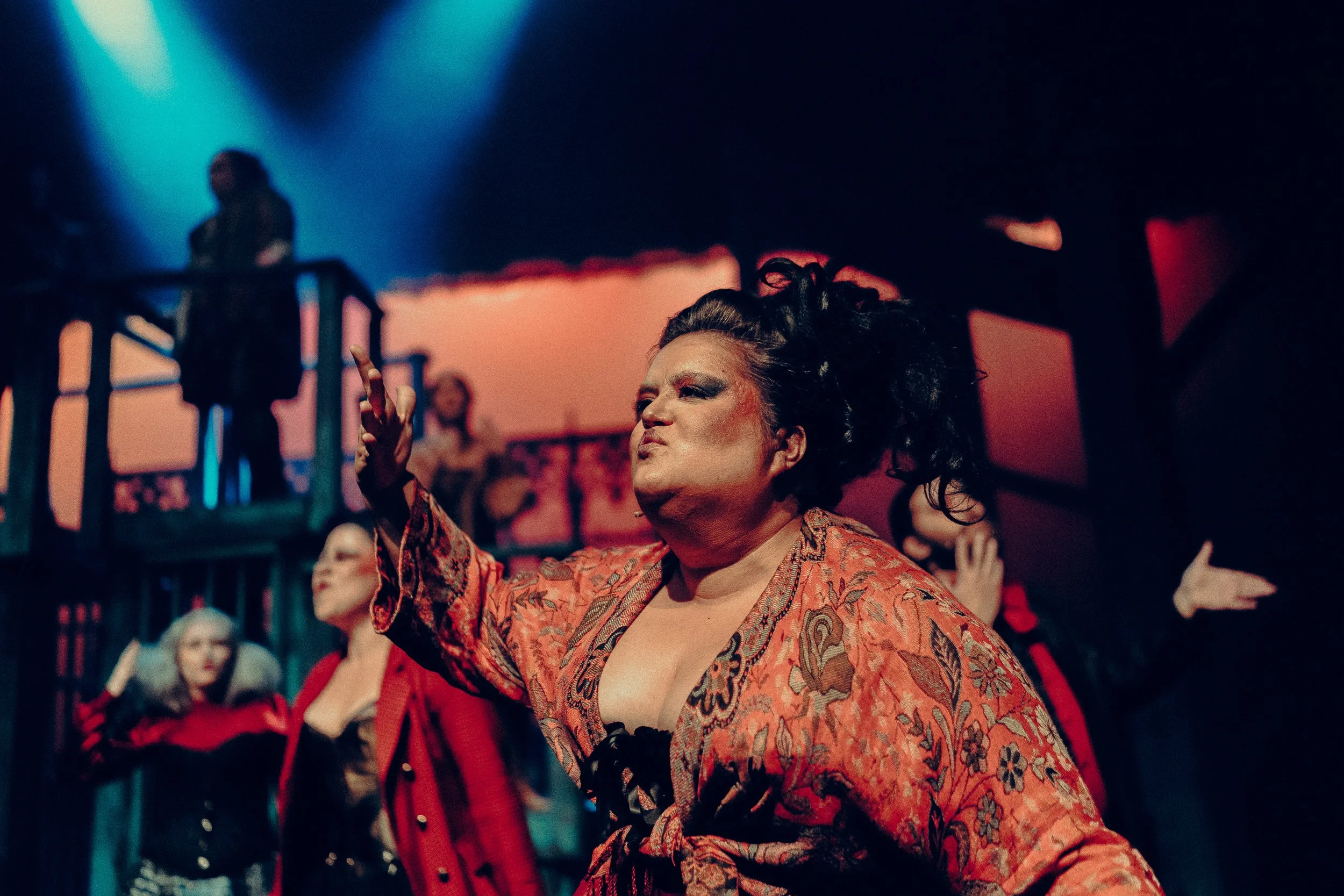 Theatrical performance with a person in dramatic makeup and costume on stage, surrounded by other performers in a colorful, dimly lit setting.